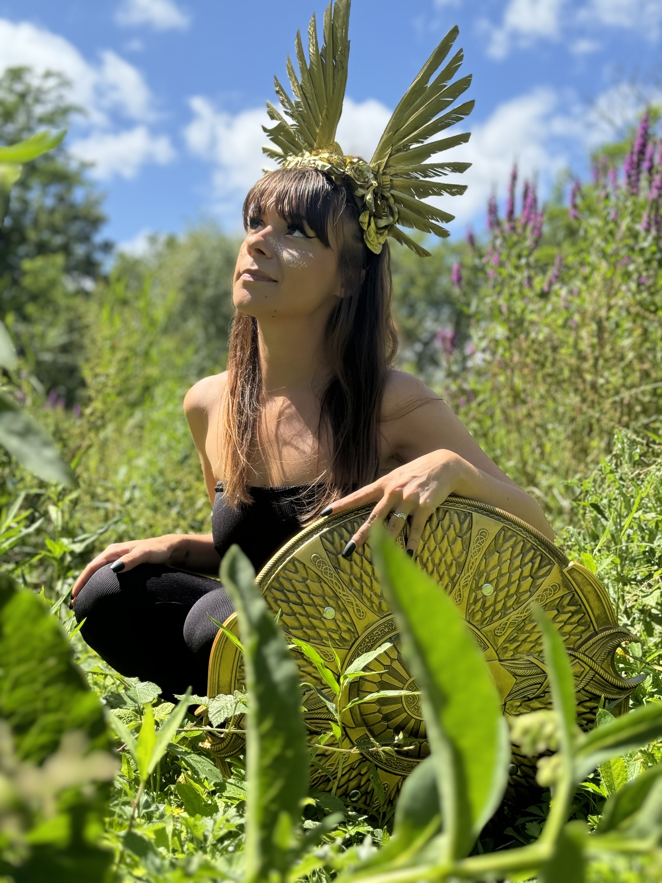 Une femme est dans une forêt tel une guerrière avec un bouclier, vêtue de noir et à genoux, regarde vers le ciel avec confiance. Ce symbole puissant d'empowerment et de sororité évoque la force de la communauté féminine et le soutien.