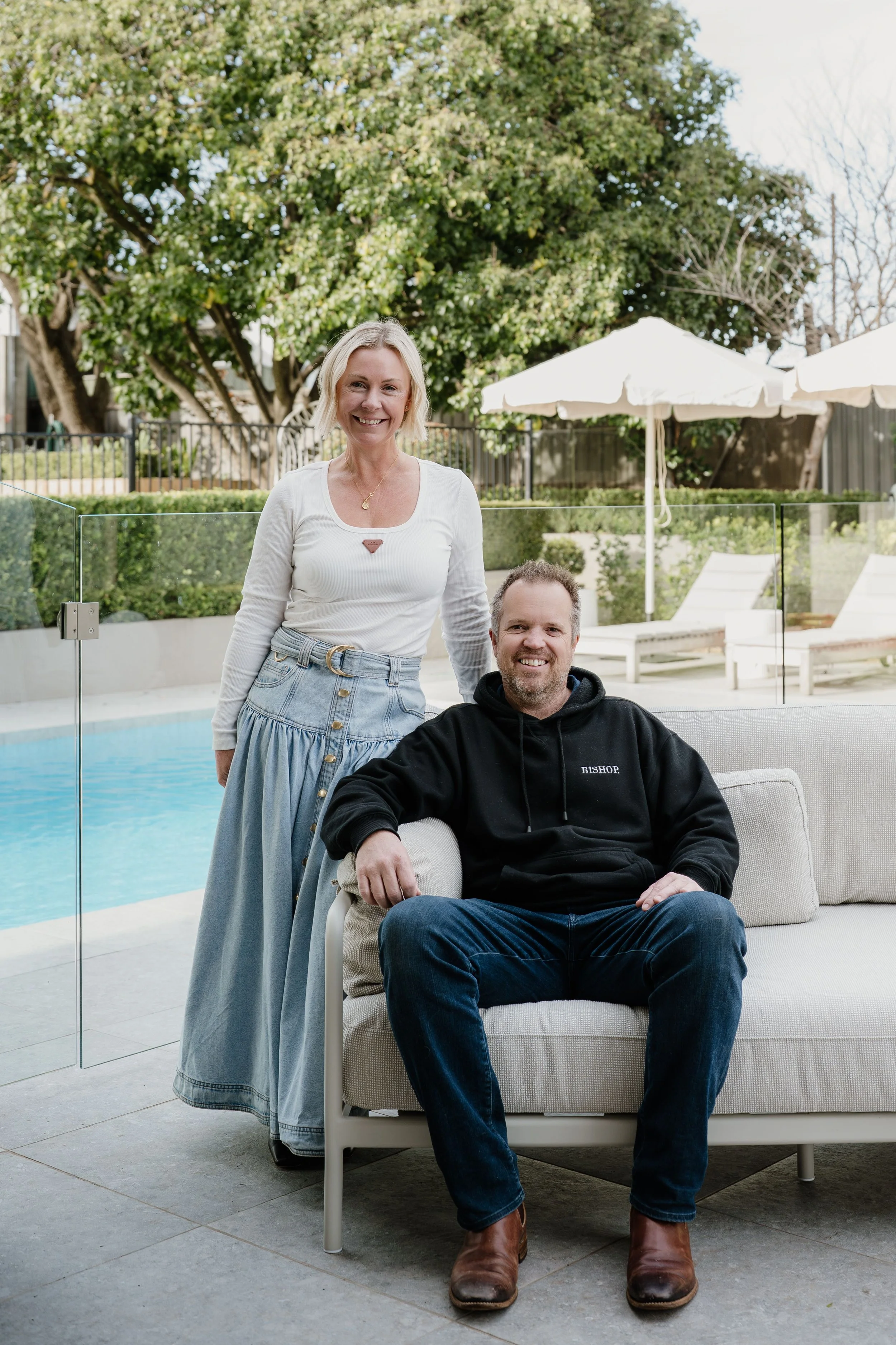 A woman and a man outdoors by a pool with umbrellas and greenery in the background. The woman is standing next to the man, who is seated on a cushioned chair, smiling at the camera with a friendly and relaxed pose.