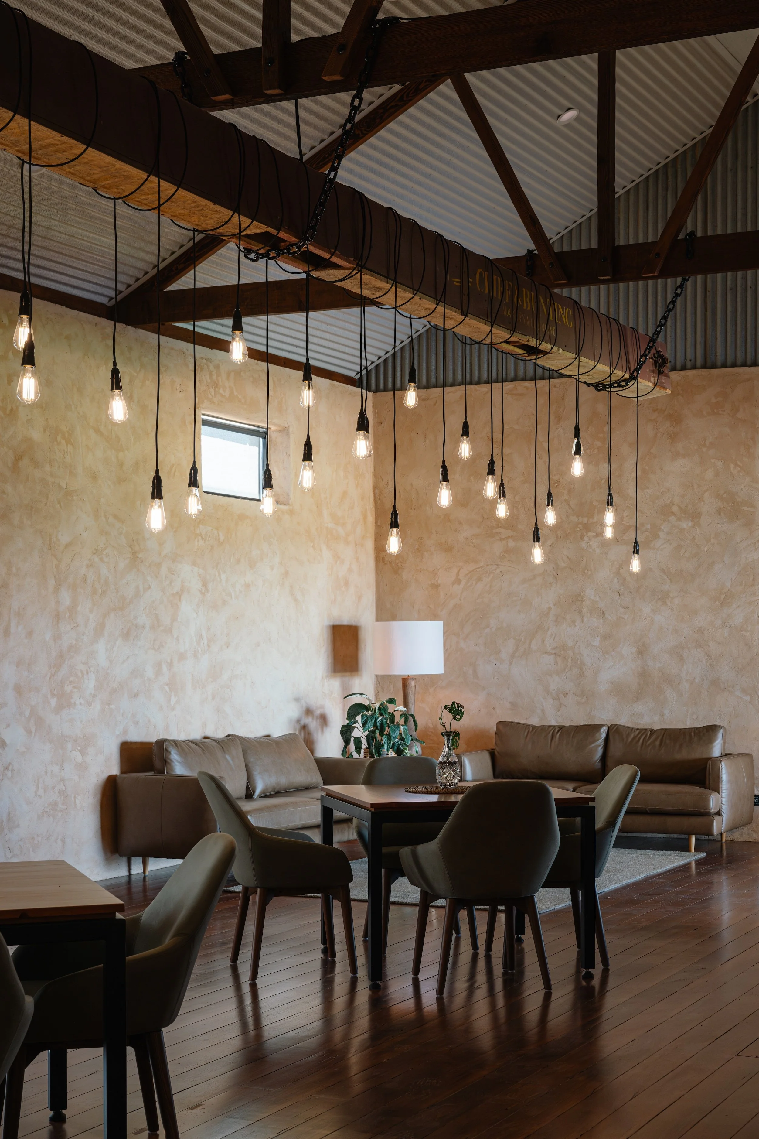 Festoon lighting suspended at staggered heights from large exposed timber beam with rustic corrugated iron ceiling detail, with casual lounge seating and dining area
