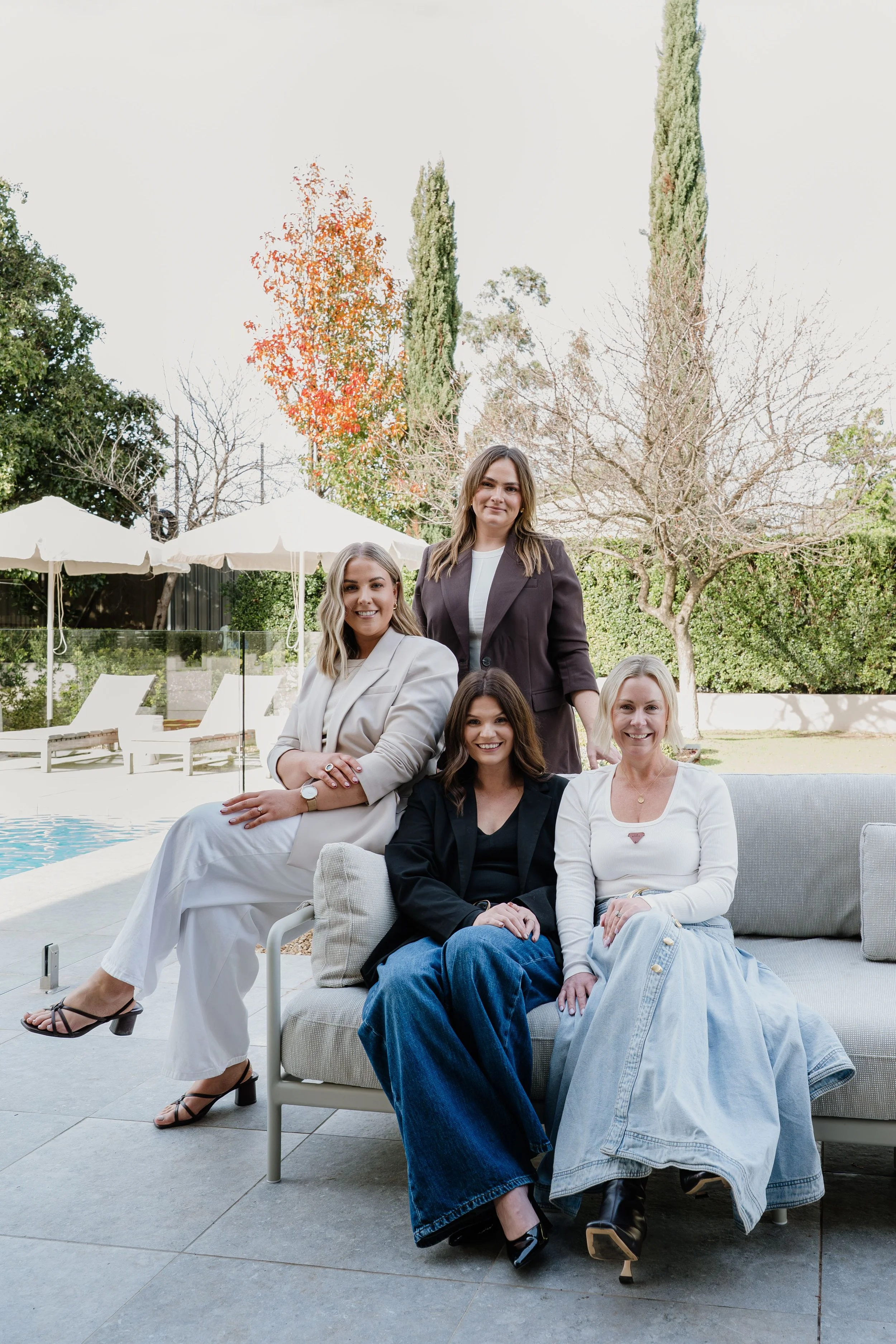 A group of four women sitting and standing outdoors near a pool, seated on a lounge chair,  with a backdrop of umbrellas, trees, and a pool in the background. Smiling at the camera with relaxed friendly poses.