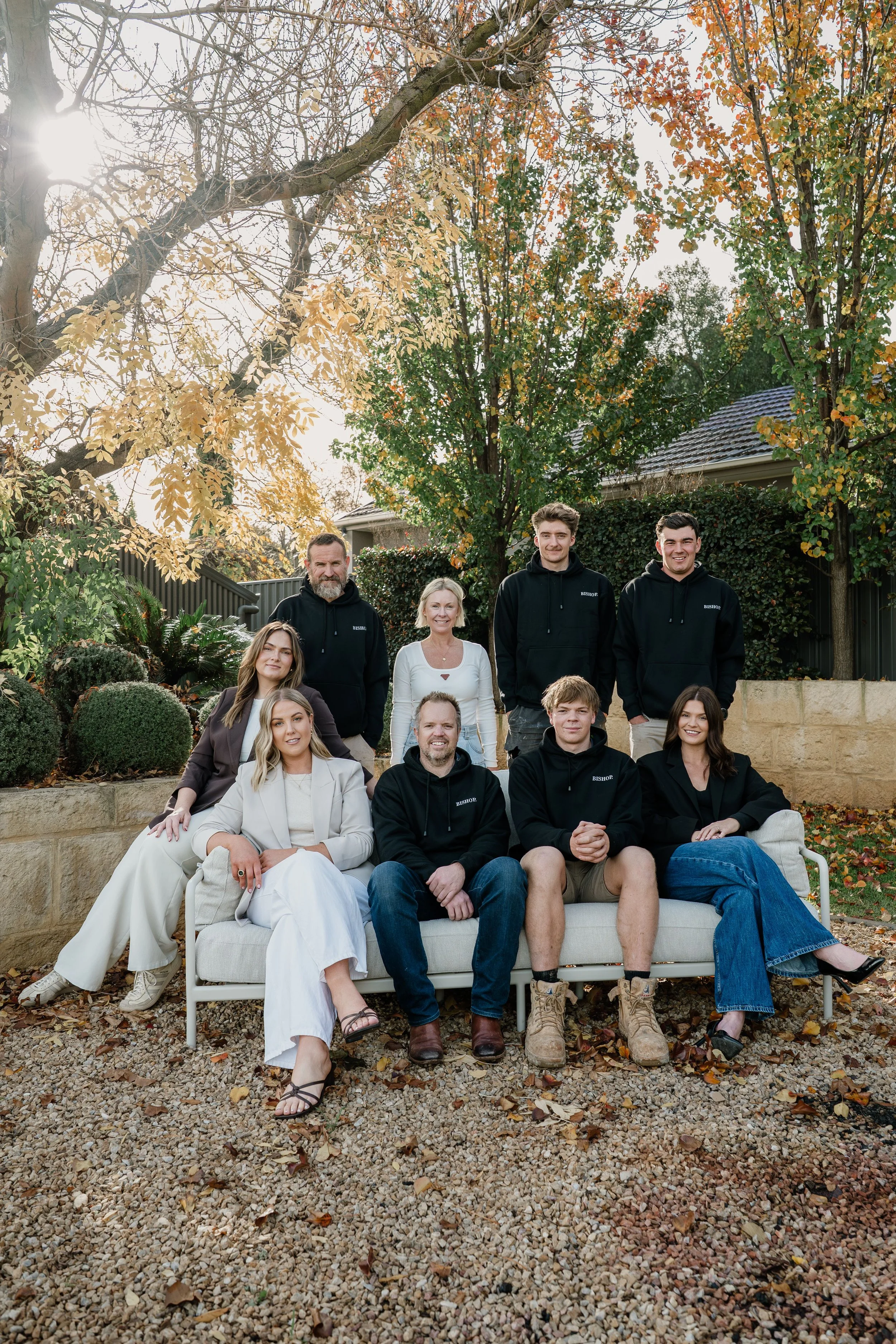 A group of nine people, five men and four women, posing outdoors in a garden during autumn with trees in the background. They are sitting and standing on a white outdoor sofa, smiling at the camera with relaxed friendly poses.