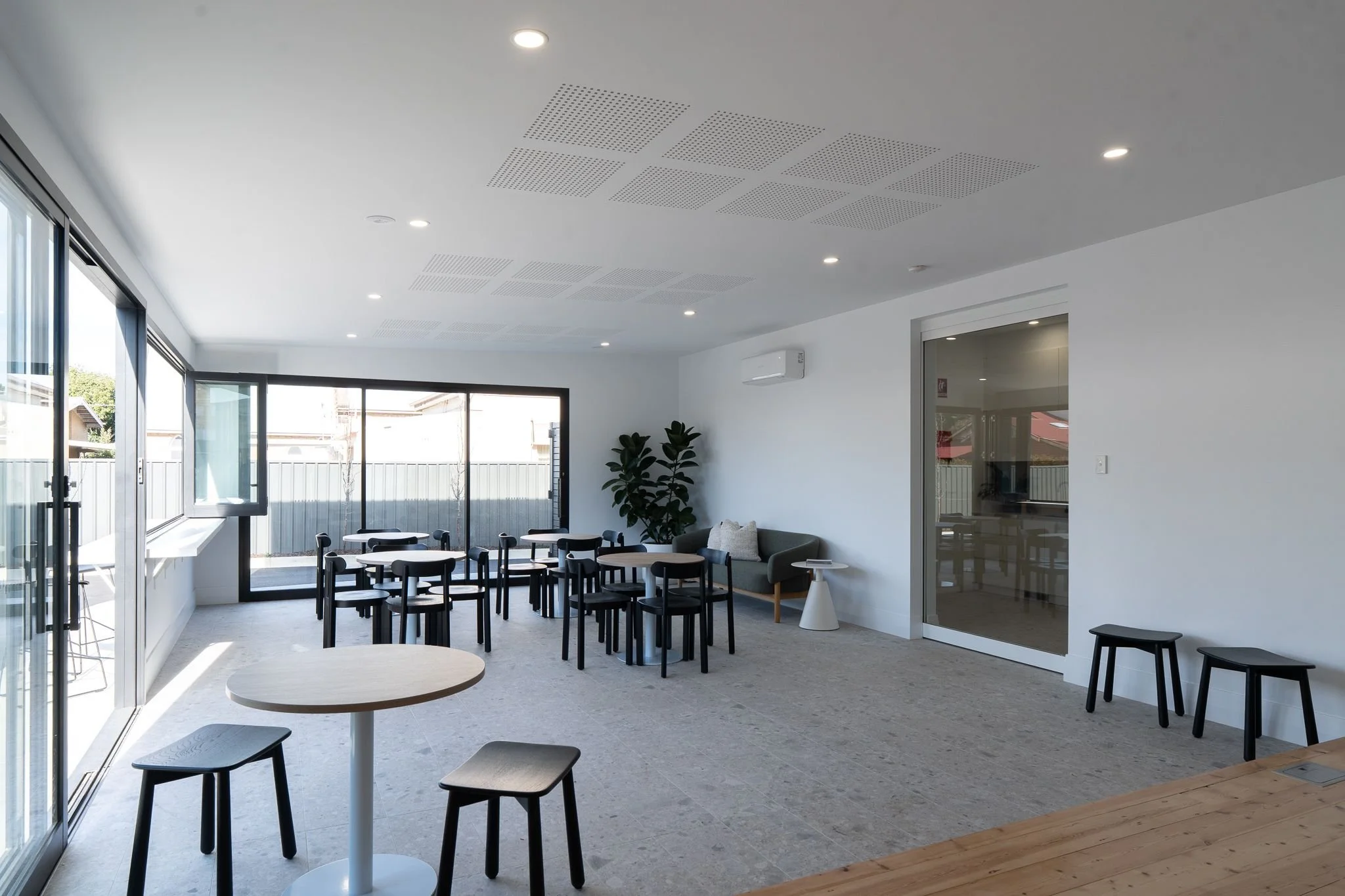 A modern, bright indoor space with ceiling lights, large windows, and a sliding glass door leading outside. There are three small tables and numerous black chairs, a sofa with cushions, and a potted plant along with a wall-mounted air conditioner.
