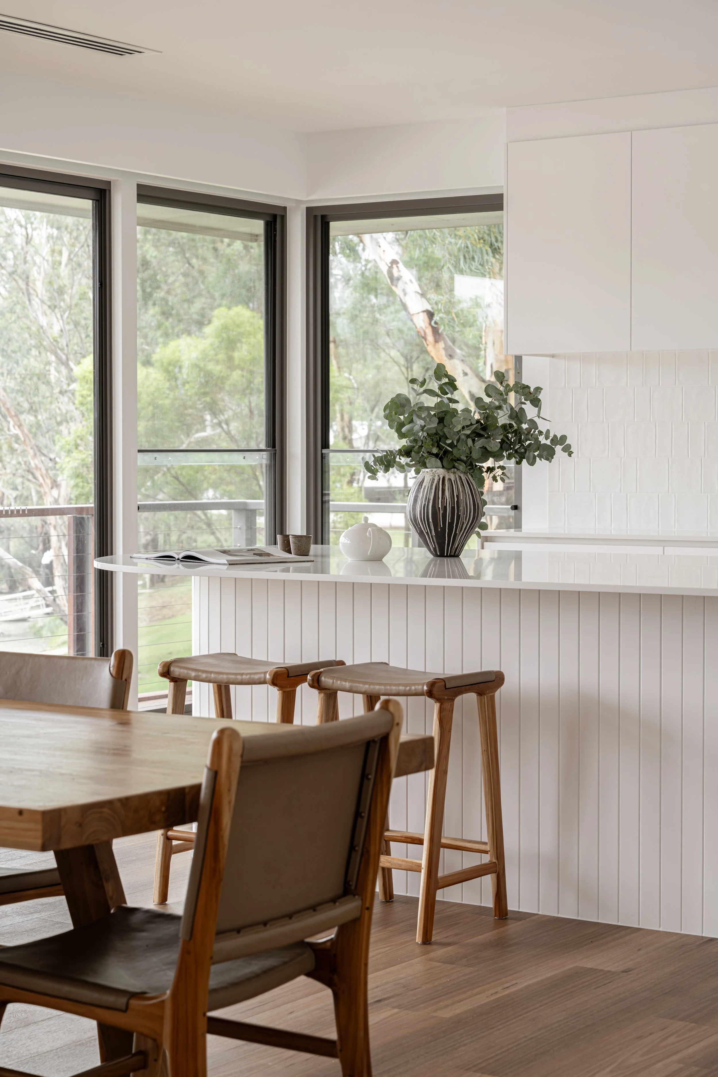A dining area with a wooden table and chairs near large windows showing trees outside. A white kitchen island with a plant and decorative items is visible.