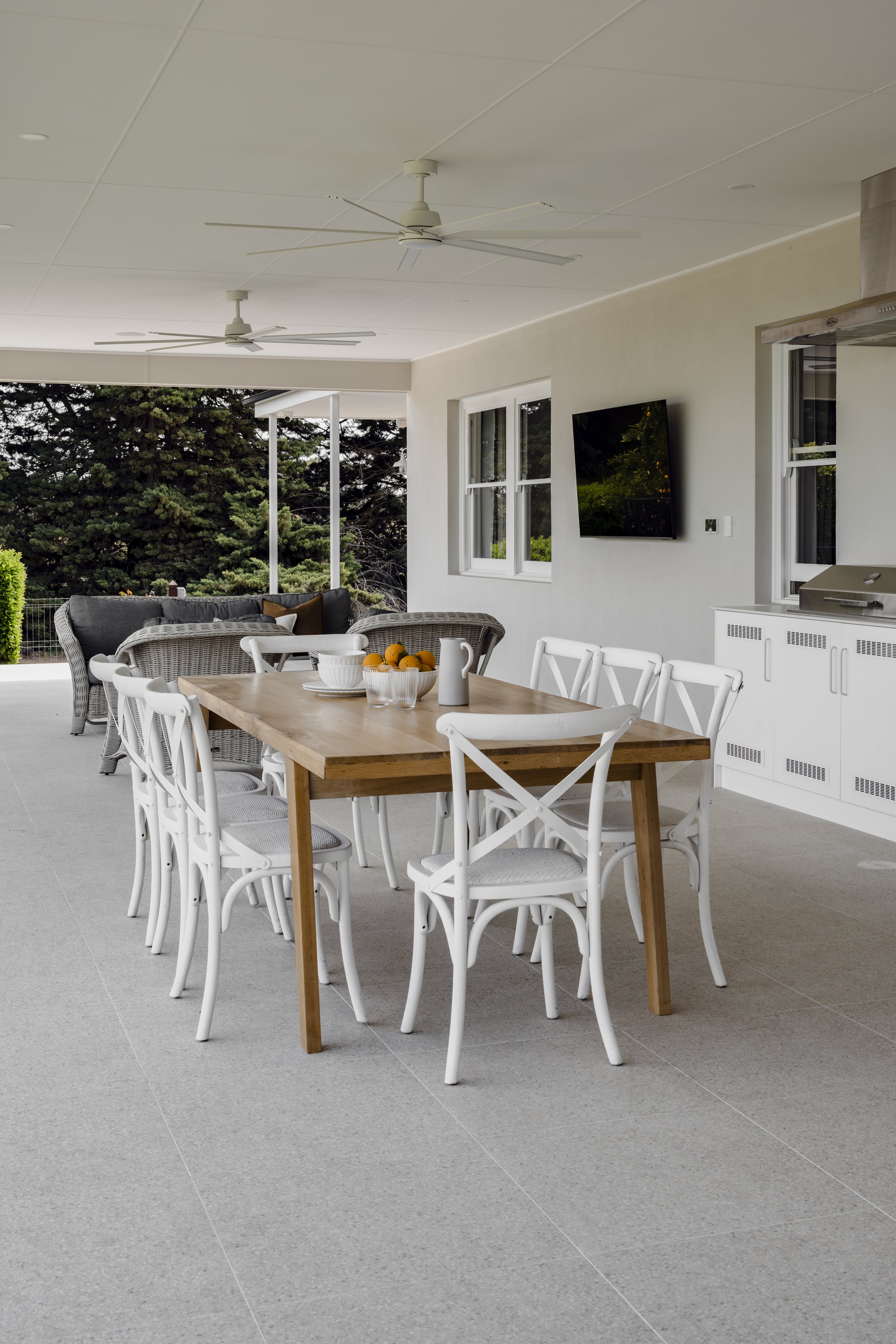 Covered outdoor patio with a large wooden dining table surrounded by white chairs, a grey wicker sofa with cushions, a mounted flat-screen TV on a white wall, ceiling fans, windows showing trees outside, and a countertop with a barbecue.