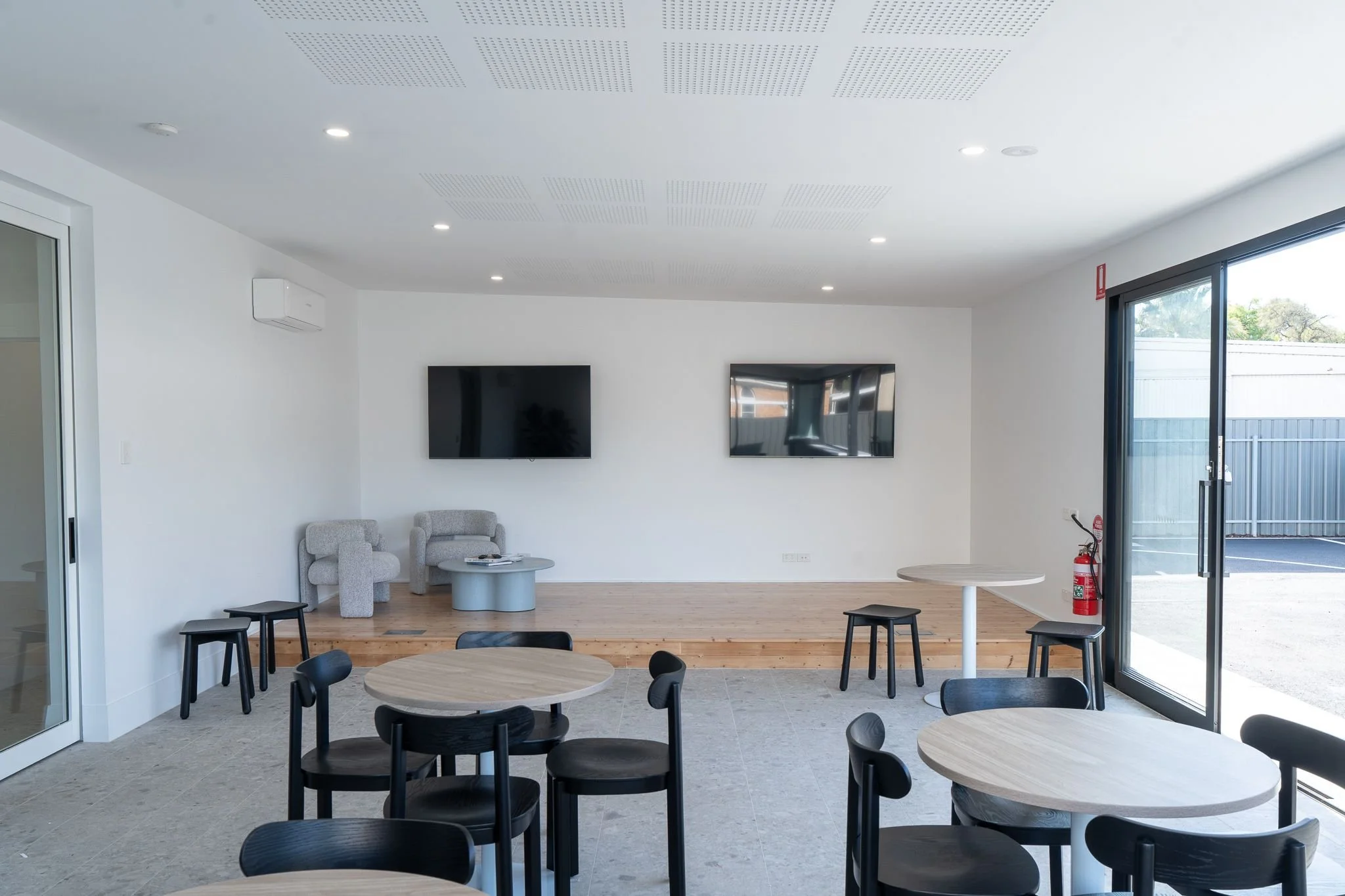 Modern lounge area with round tables and black chairs, TV screens on white wall, large glass sliding door to outside, and wooden raised platform with modern grey chairs around several round tables.