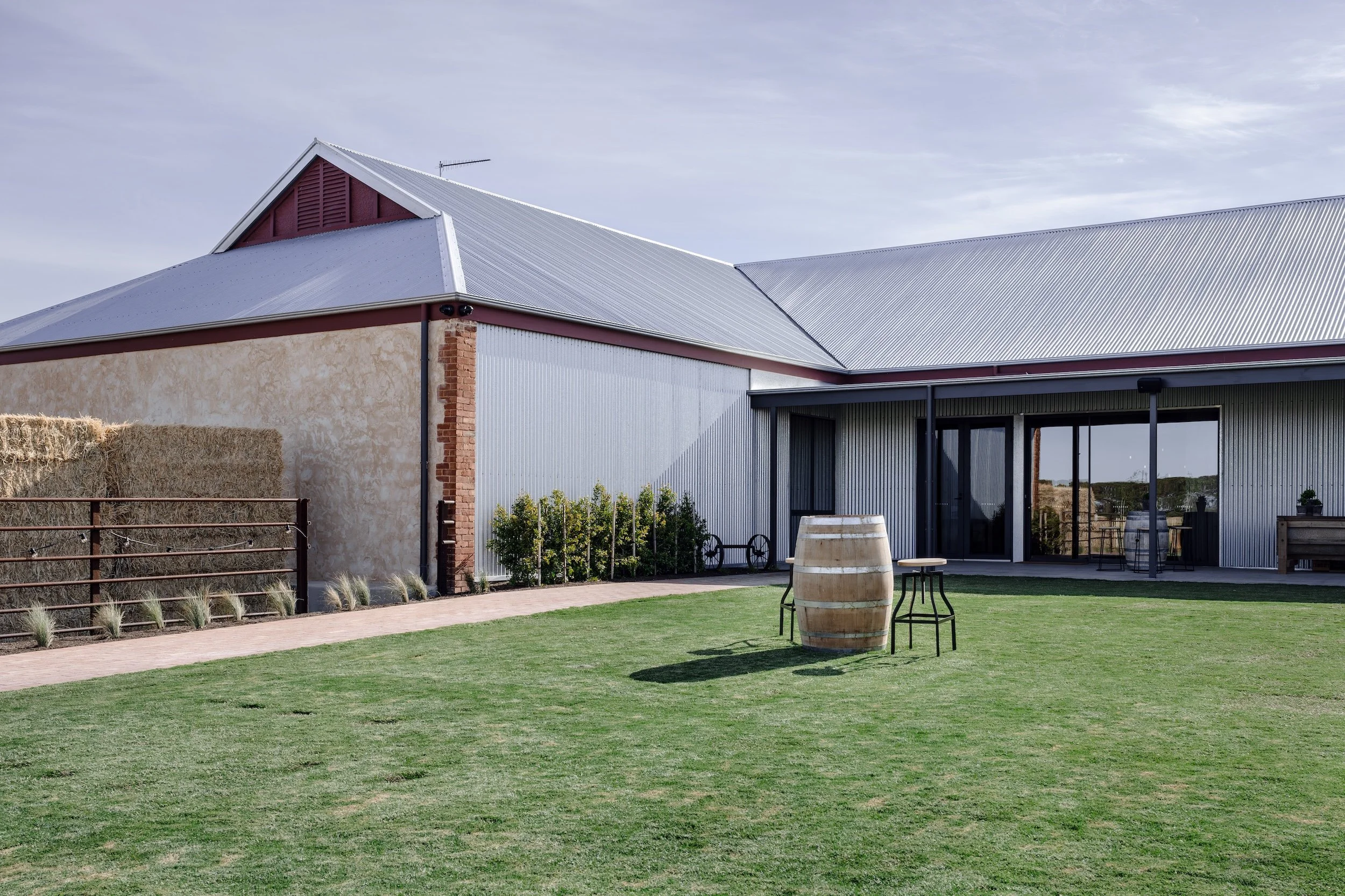 Restored barn exterior with barrel table on lawn, hay bales behind rustic rail fencing and native grass plantings, Yorke Peninsula