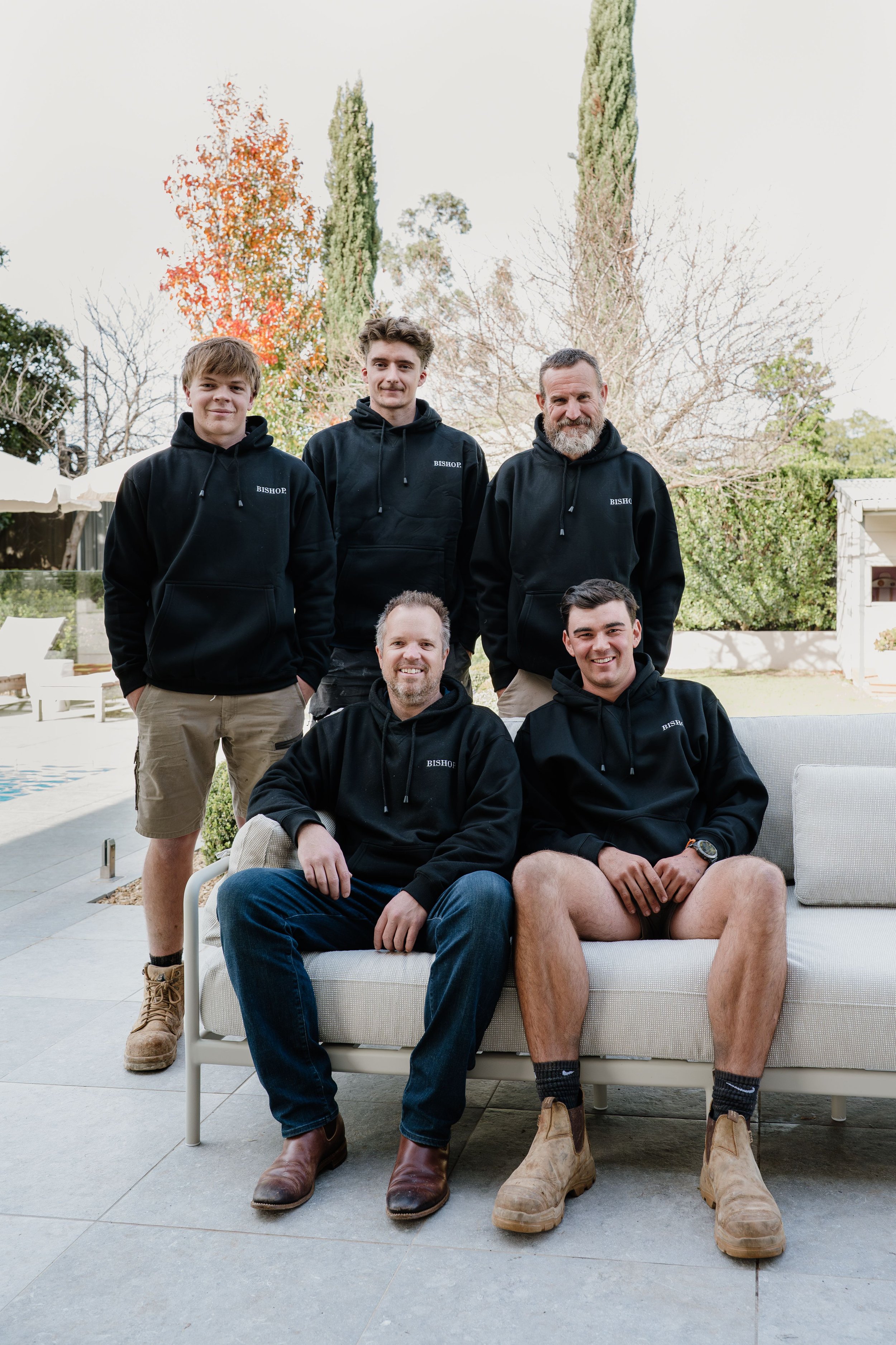 Five men in black hoodies with the word 'BISHOP' standing and sitting outdoors in a backyard with trees, some with autumn leaves, and patio furniture.