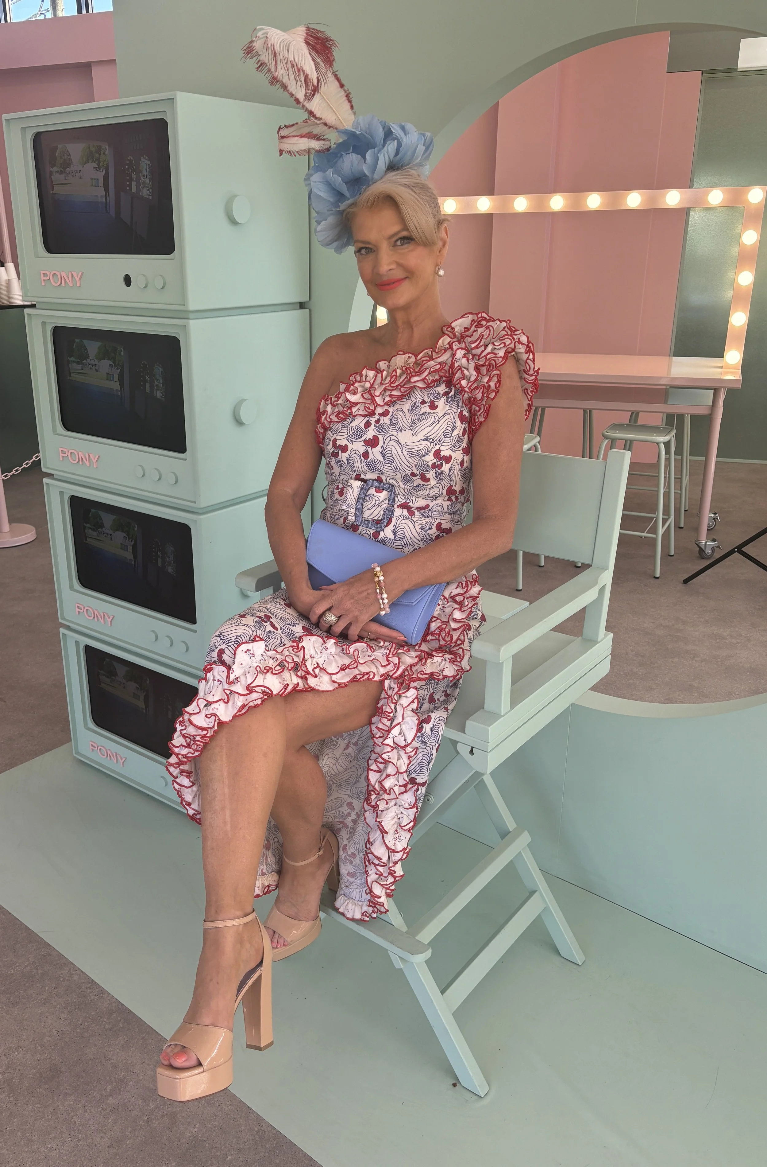 A woman sitting on a light blue bench in front of vintage TV sets with the word "PONY" on them, wearing a floral dress with ruffled shoulder details, beige platform shoes, a large blue hat with feathers, holding a light blue clutch, and standing in an indoor space with pastel-colored walls and bright lighting.