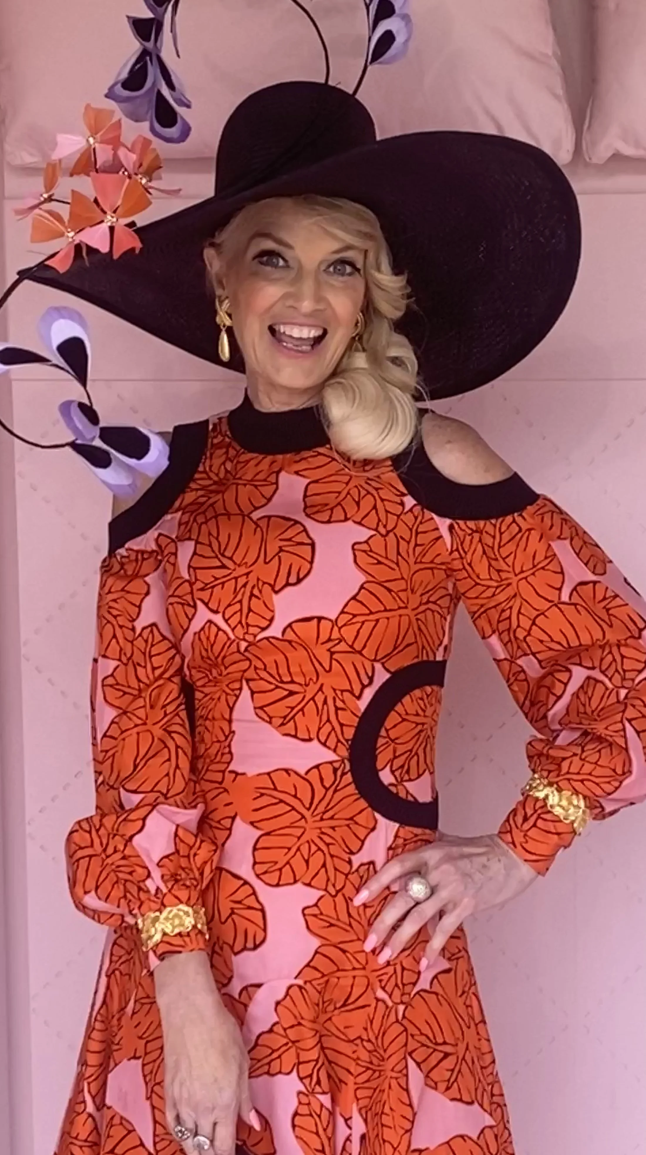 A smiling woman wearing a large black hat with decorative purple and pink bows, gold earrings, and a colorful dress with a leaf pattern in shades of orange and pink. She is standing in front of a pink background with valentine-themed decorations.
