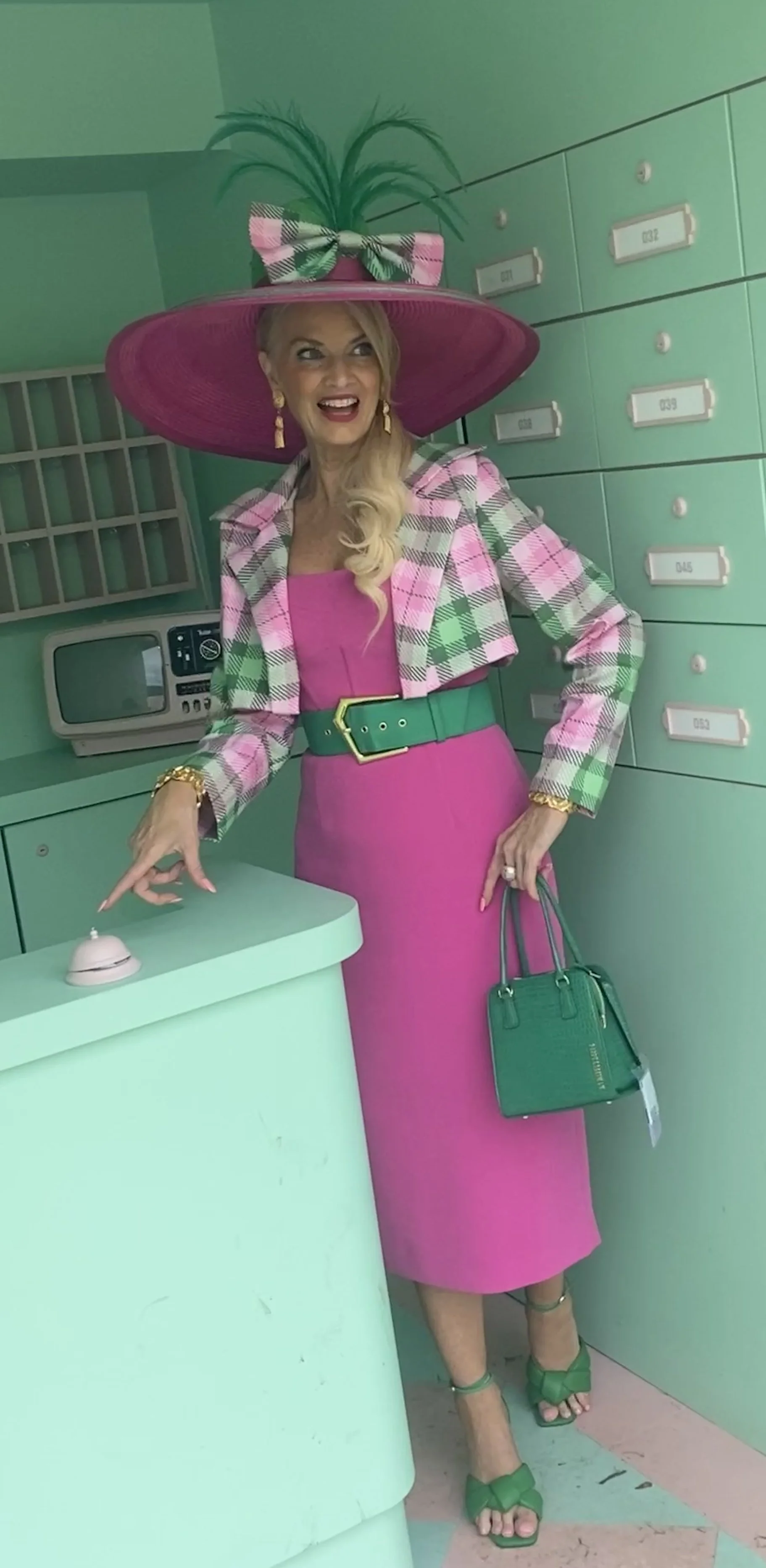 A woman in a pink dress, plaid blazer, and wide-brimmed pink hat with a bow and feathers, standing at a counter inside a retro-style mint green building. She is smiling, holding a small handbag, and pointing at a bell on the counter.
