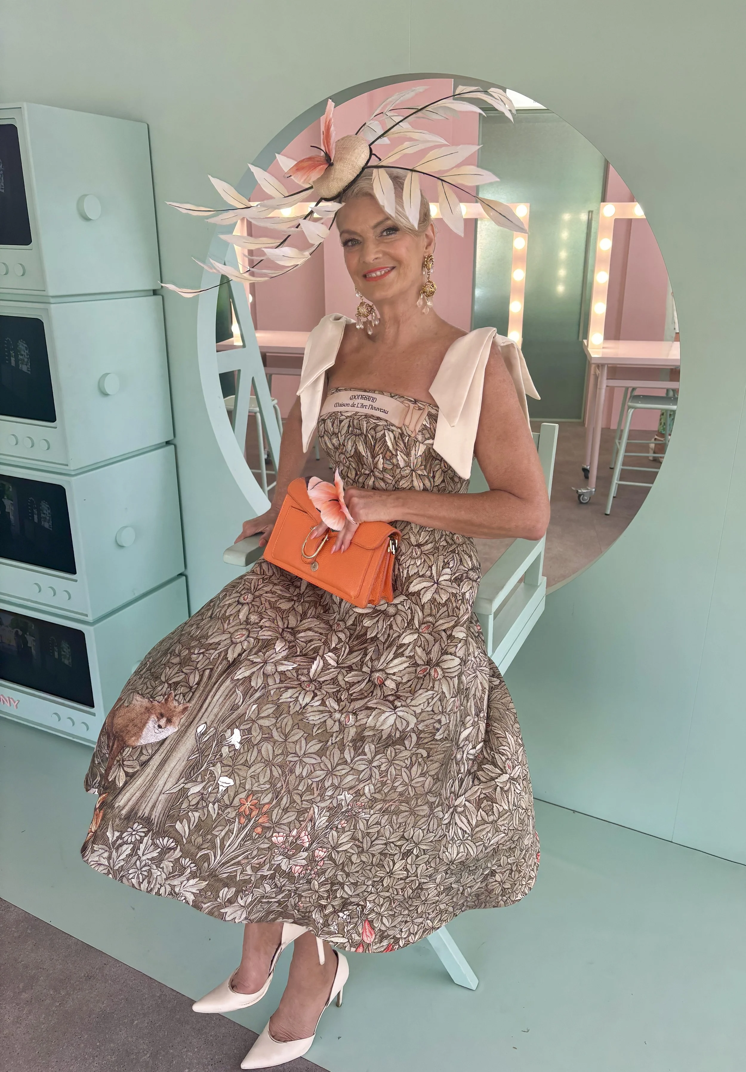 Statement millinery in neatural tones with feathers, ostrich quills, to give a botanical leaf and forest feel. Worn with a floral dress, white heels and orange handbag in a colourful dressing room.