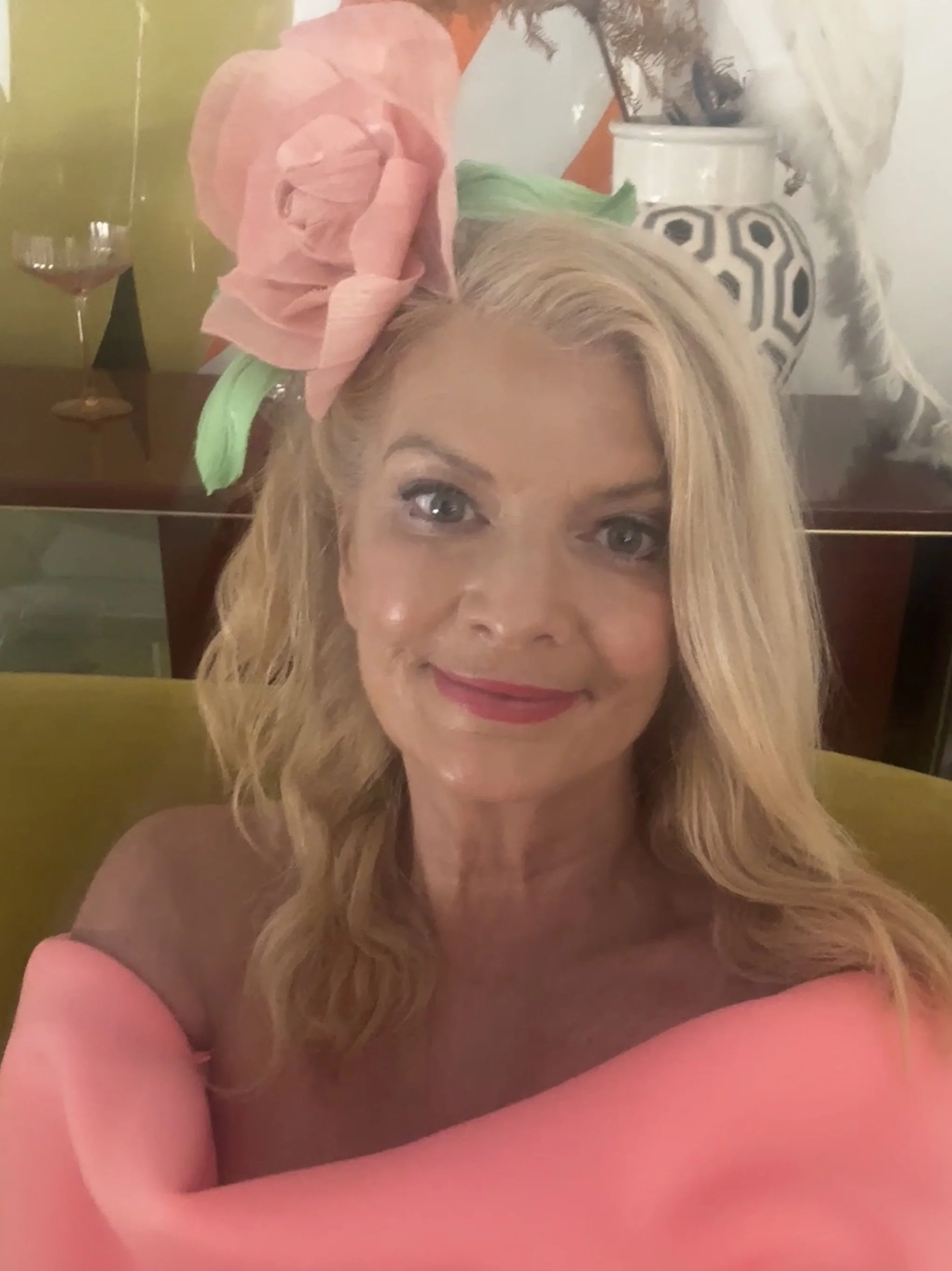 A woman with long blonde hair and light skin, smiling, wearing a pink top, with a large pink and green paper flower on her head. In the background, there is a wall painted green, a dark wooden table with a glass of pink liquid, and decorative items including a vase with black and white patterns.