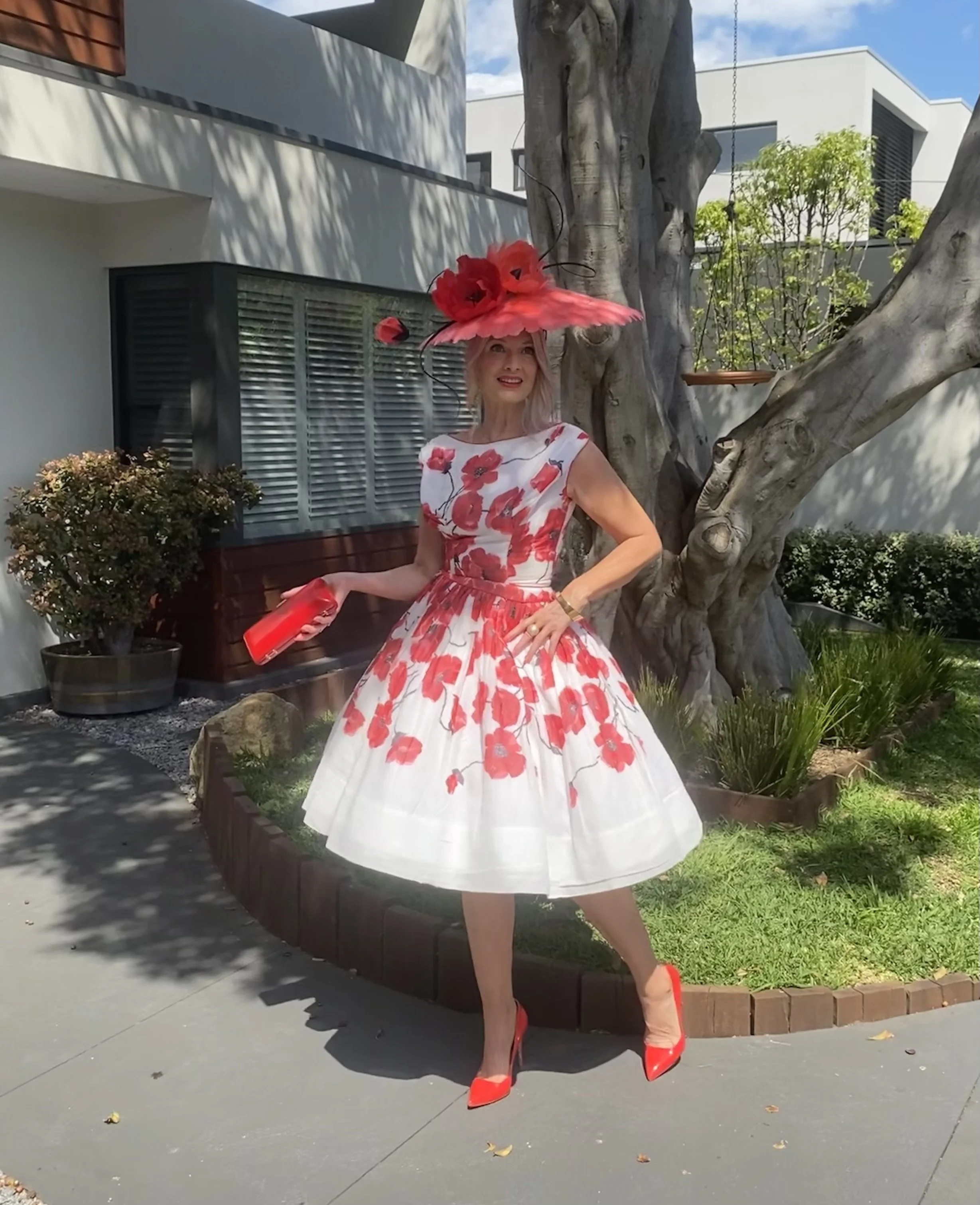 A woman wearing a floral dress, a large bright pink hat with flowers, and red high heels stands outdoors near a tree and a modern house with white walls and black window shutters.