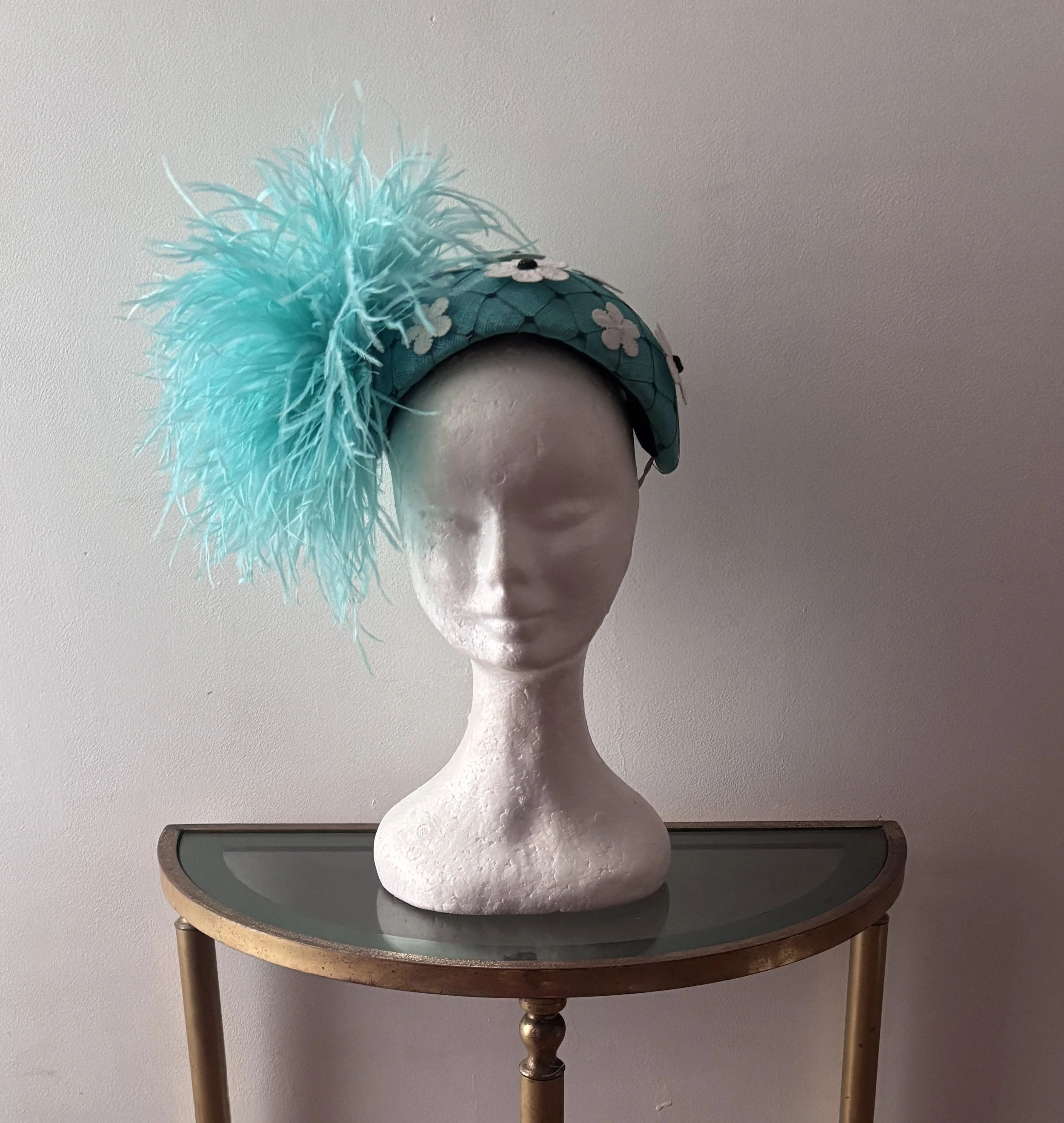 A mannequin head on a table wearing a blue hat with white flowers and a large light blue feather decoration.