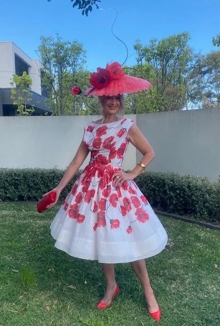 Statement coral red millinery hat with hand-made feather poppy flowers and black ostrich quills, showcasing sculptural handcrafted headwear, worn with a white and red floral poppy dress by a smiling woman outdoors.