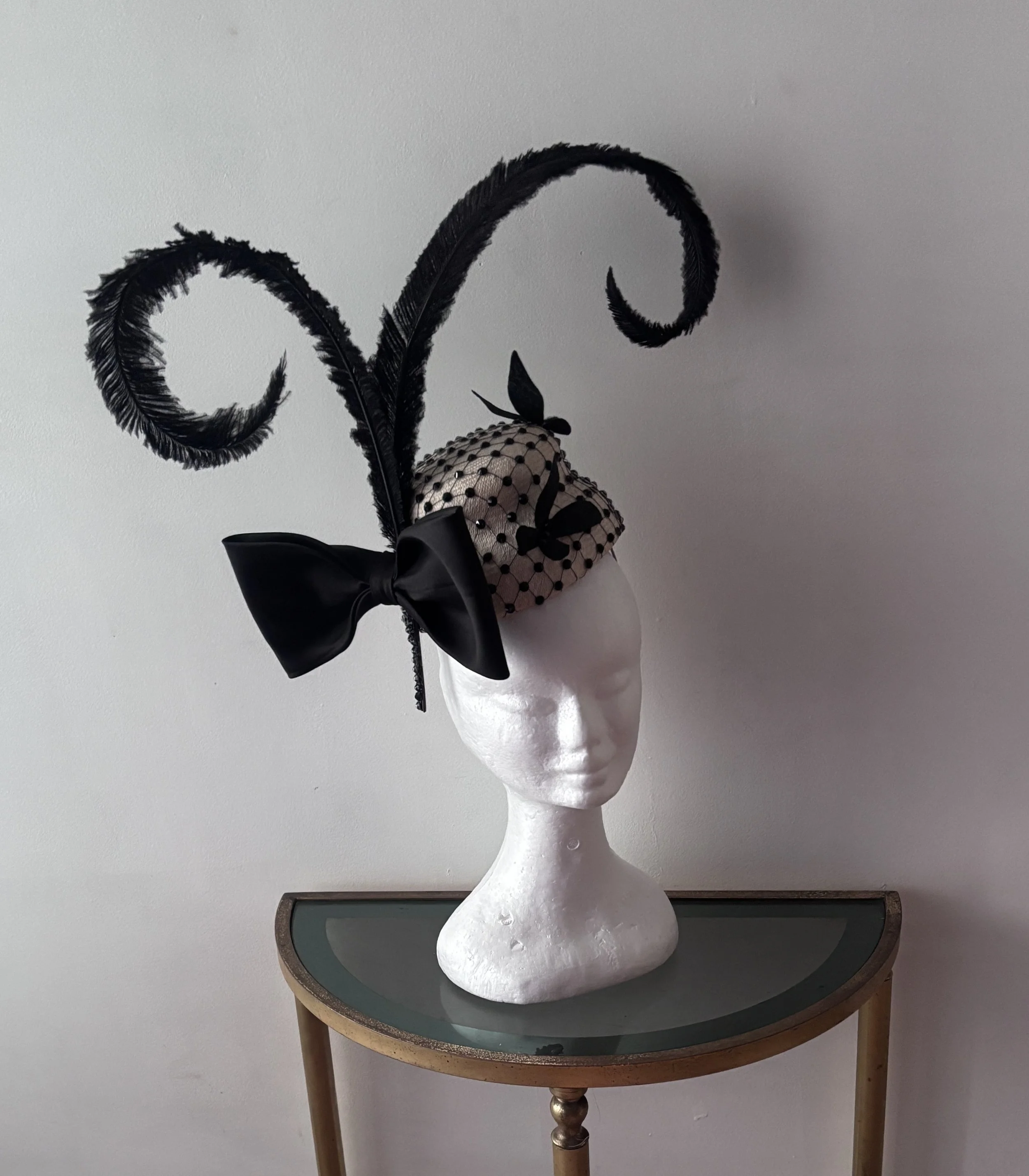 A mannequin head with no facial features wearing an ornate costume headpiece on a green table with wooden legs against a plain white wall.