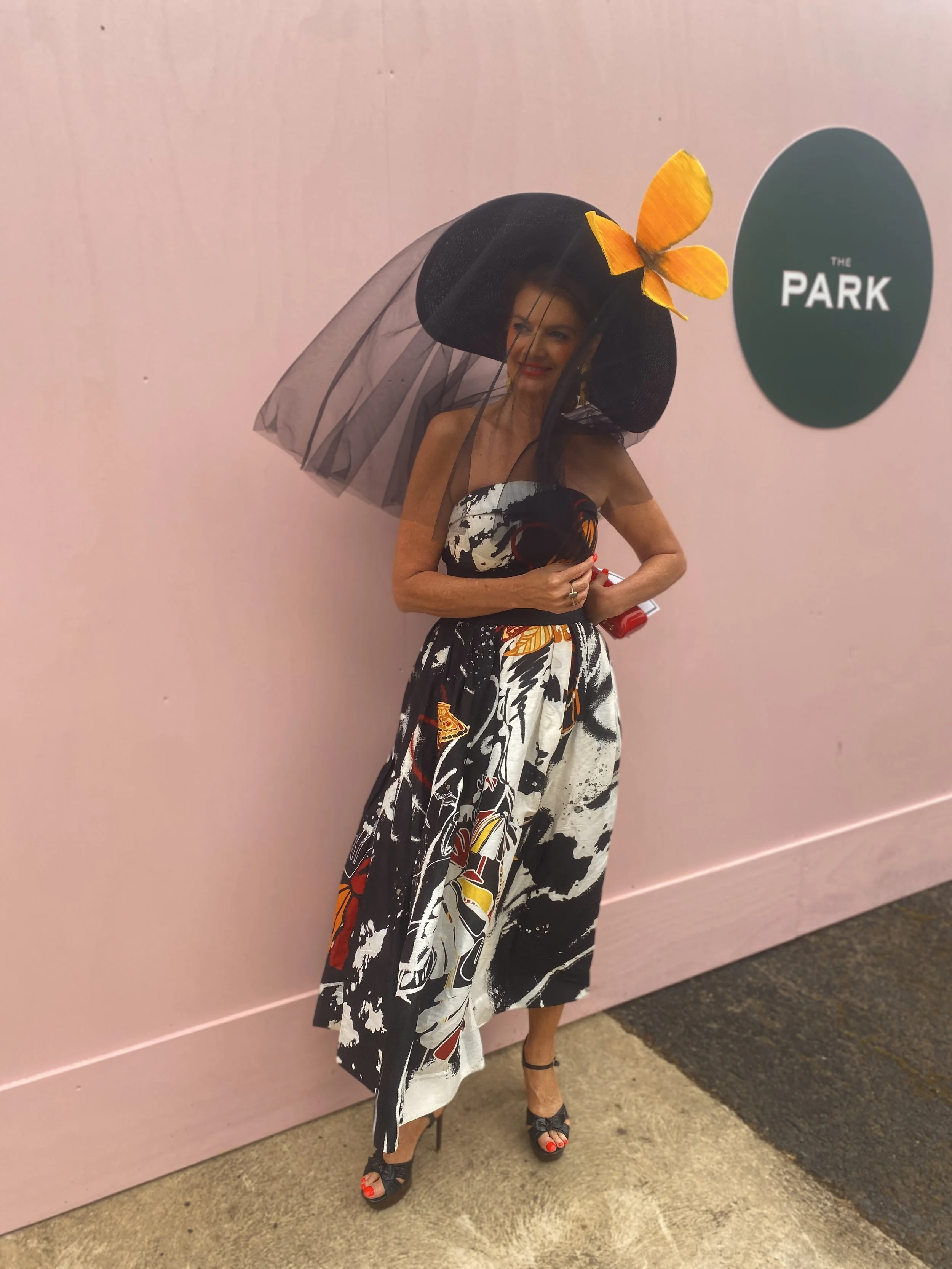 A woman in a strapless dress with a black and white abstract pattern and red accents, wearing a large black hat with orange butterfly decoration and black veil, standing against a pink wall.
