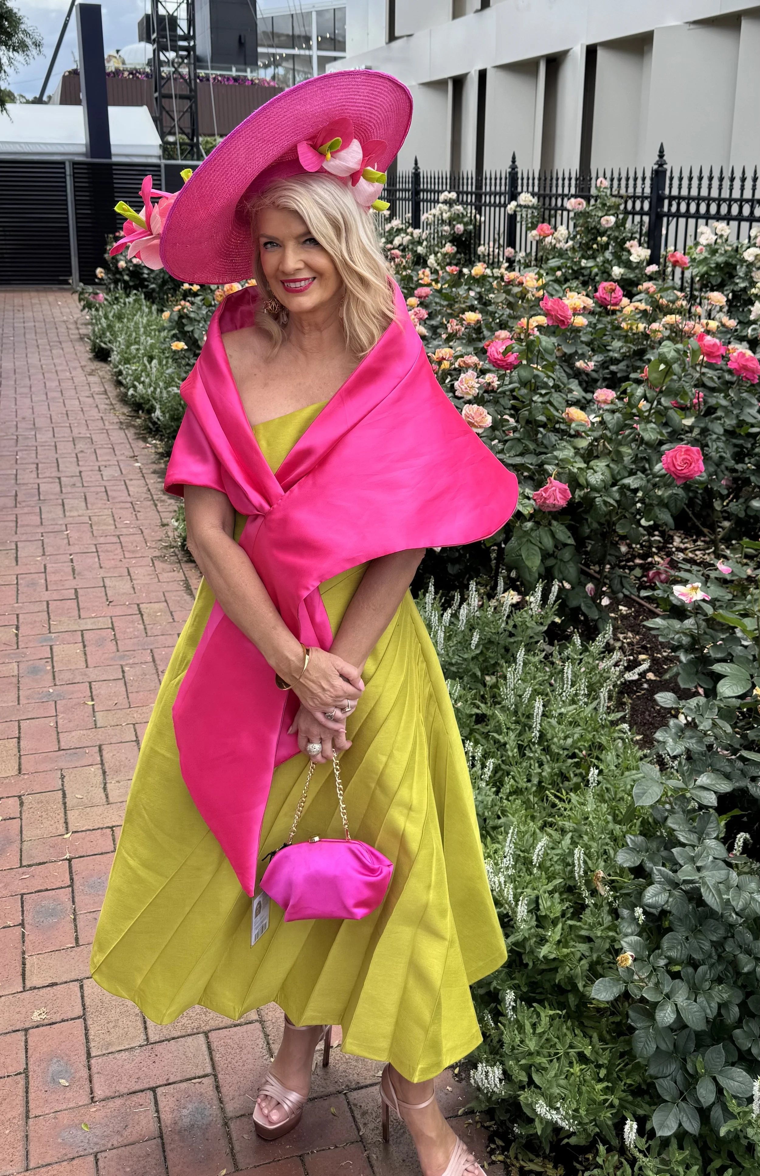 A woman standing outdoors in front of a flower garden, wearing a wide-brimmed pink hat decorated with large pink flowers, a vibrant pink shawl, and a yellow dress. She is holding a small pink purse and wearing nude high-heeled shoes, smiling at the camera.