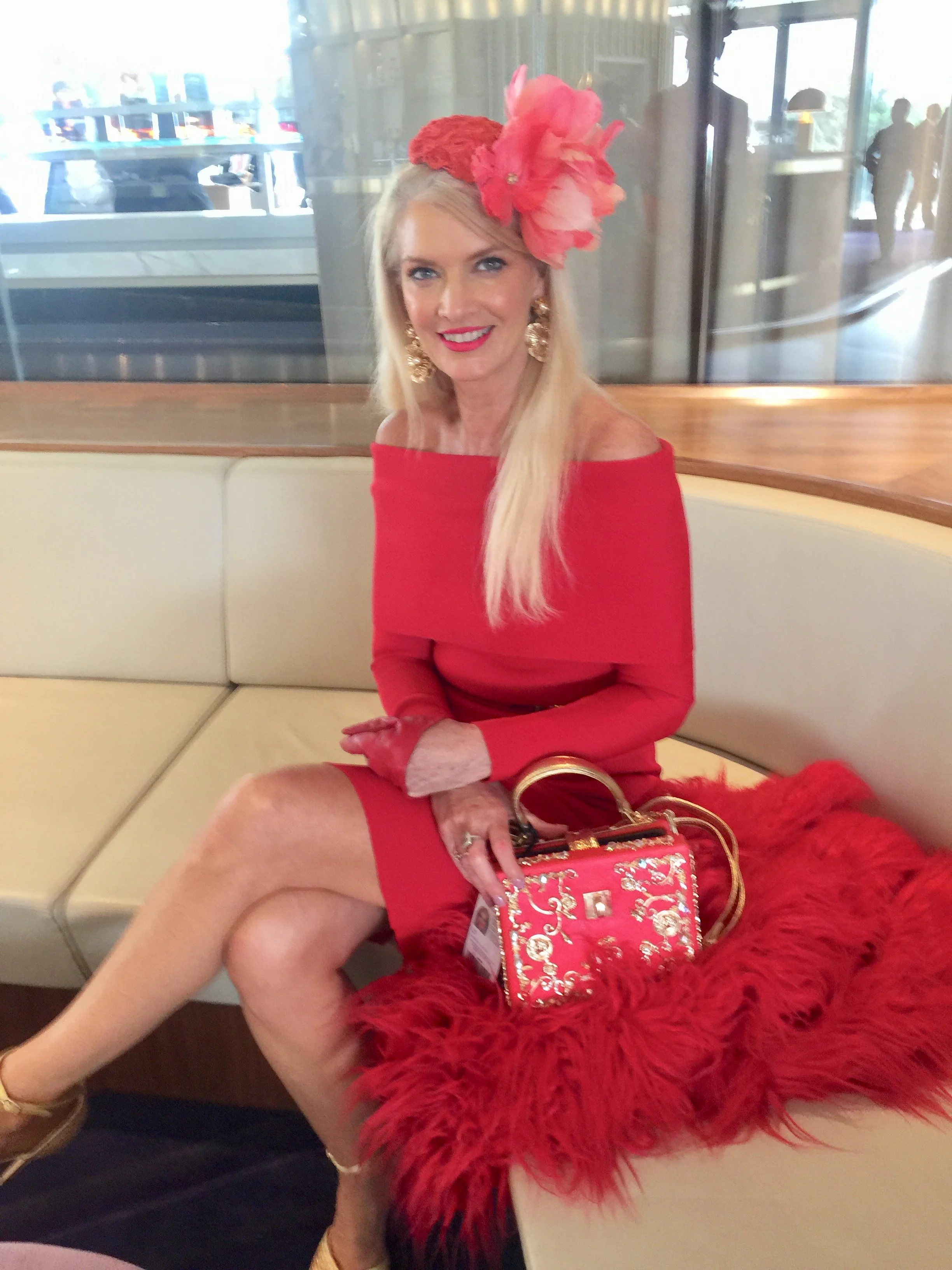 A woman sitting on a beige booth seat in a restaurant, dressed in a red off-the-shoulder dress with large gold earrings, a floral red hat, and holding a red purse, with a red feathered accessory in front of her.