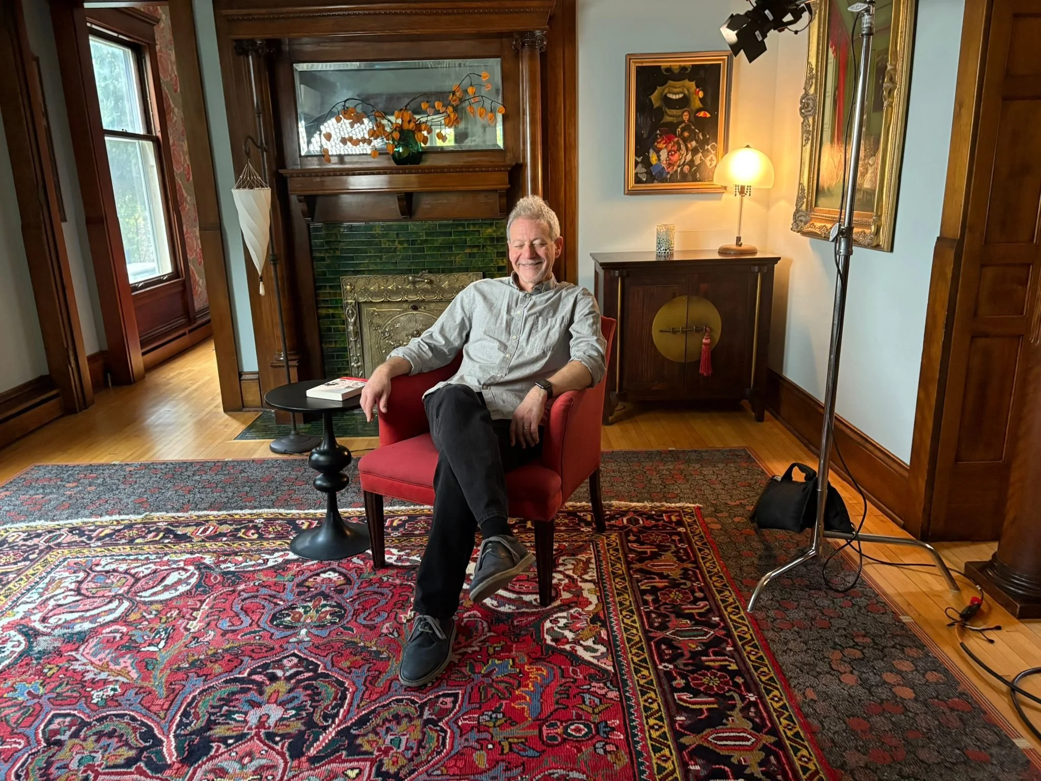 A man sitting in a red armchair in a cozy, well-decorated living room with a fireplace, colorful artwork, a side table with a book, and a studio light on a stand.