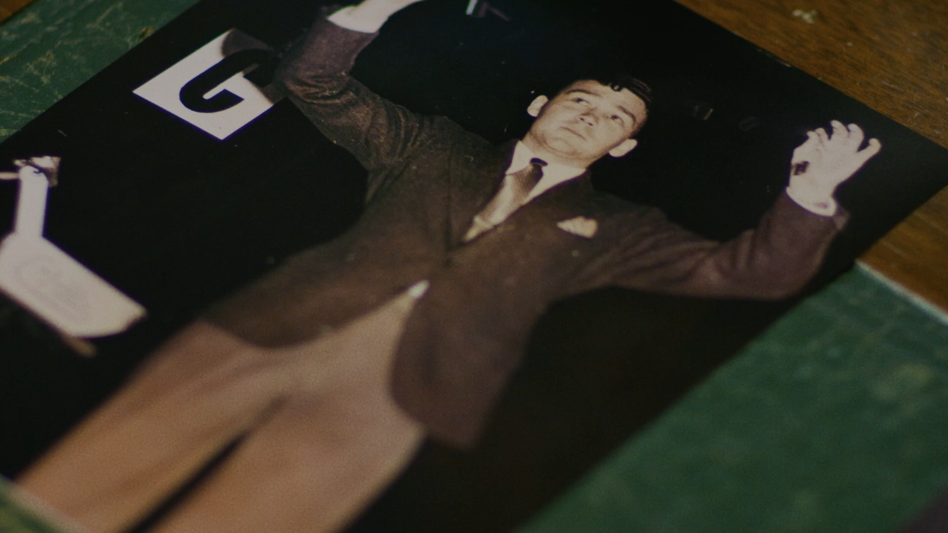 Black and white photograph of a young man in a suit with arms raised, lying on the ground with a dark background.