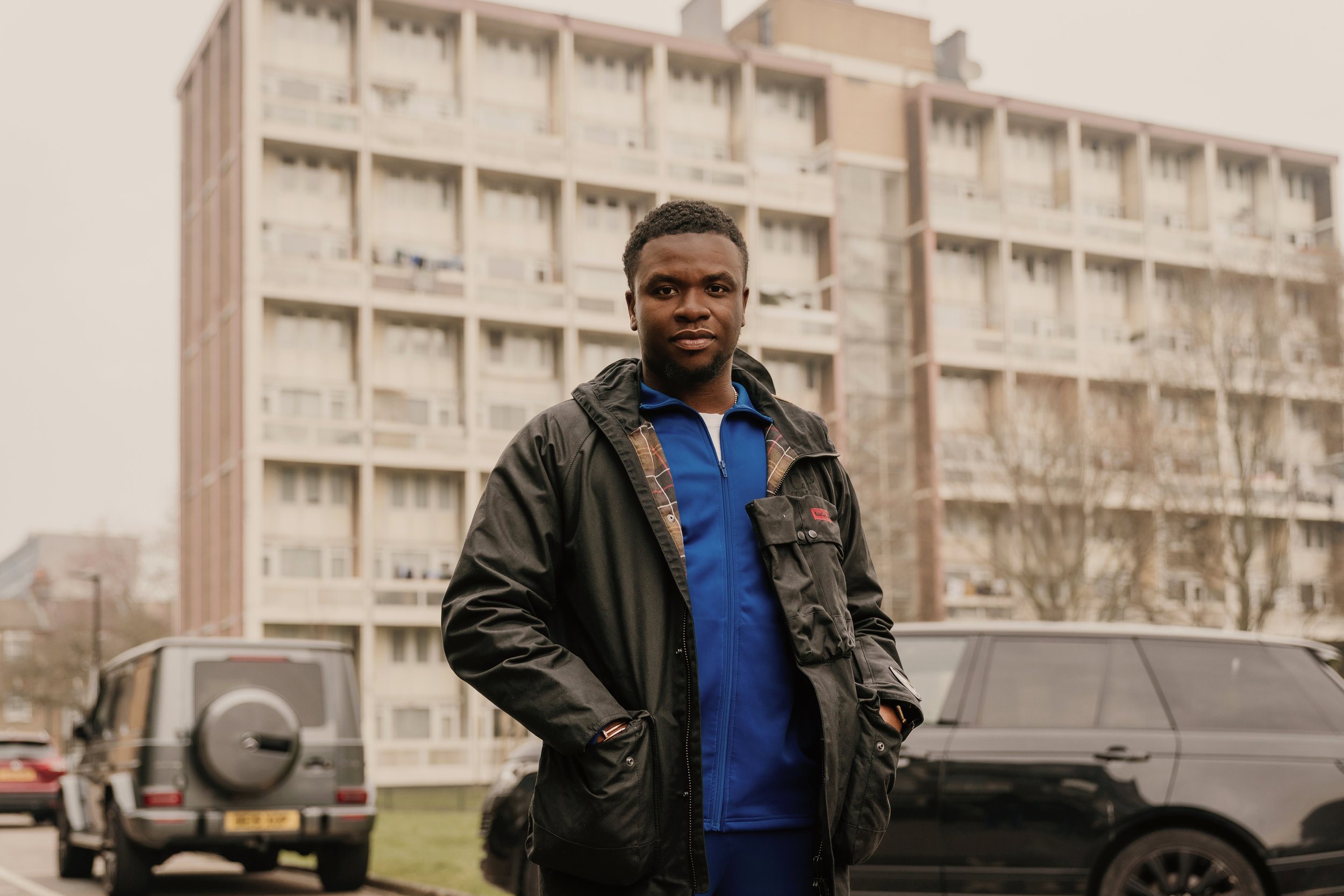A young man in a black jacket and blue sportswear standing outdoors in front of a large apartment building, with cars parked in the background.