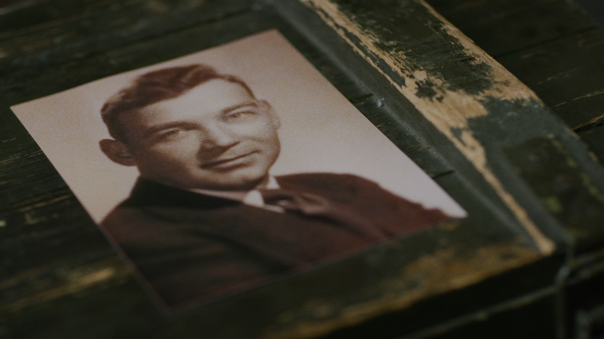 Black and white portrait photograph of a young man in formal attire, smiling slightly, placed on a weathered wooden surface.