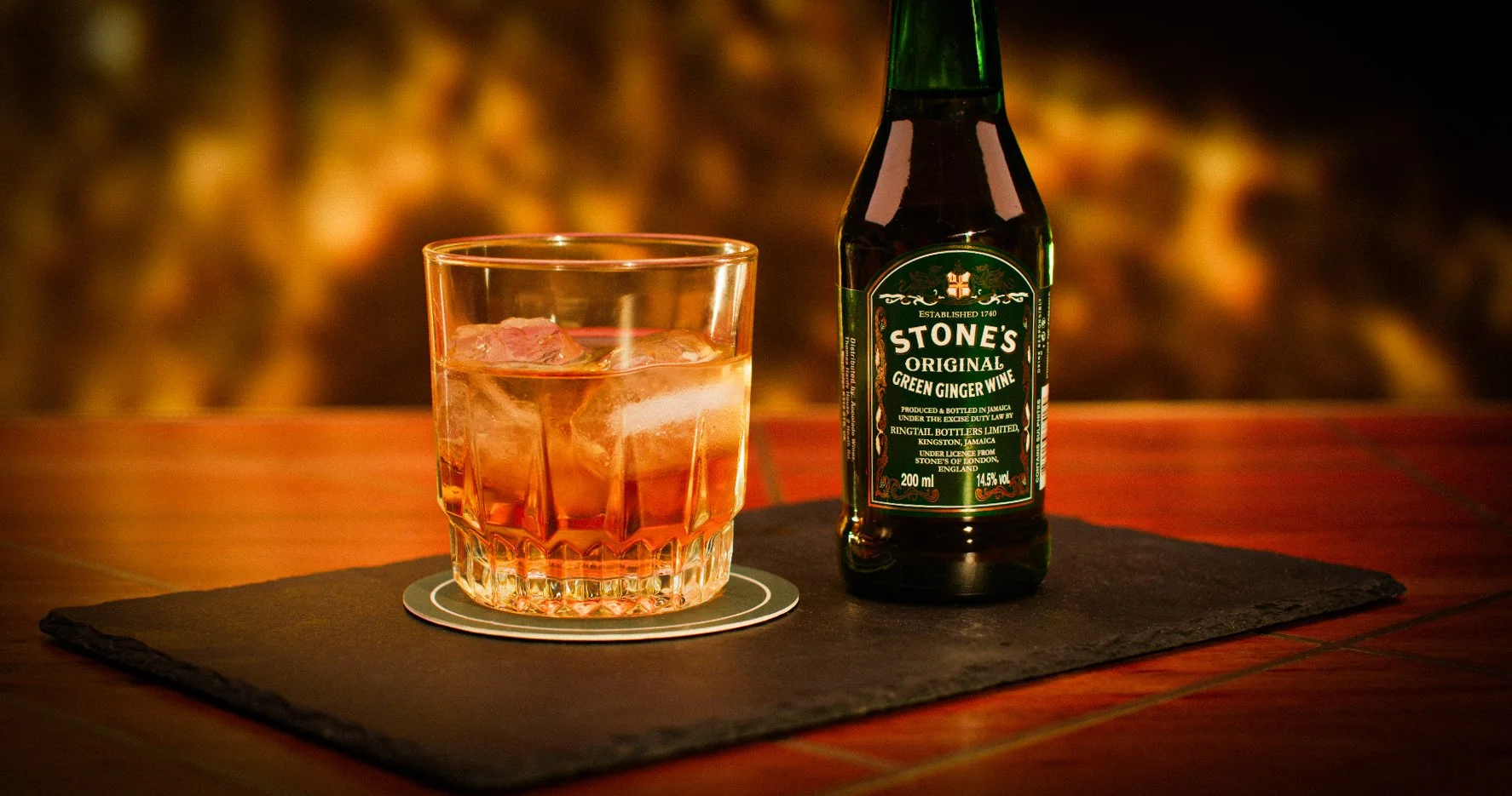 A glass of pink-colored beverage with ice cubes and a bottle of Stone's Original Green Ginger Wine on a black mat on a wooden surface, with a blurred warm-toned background.