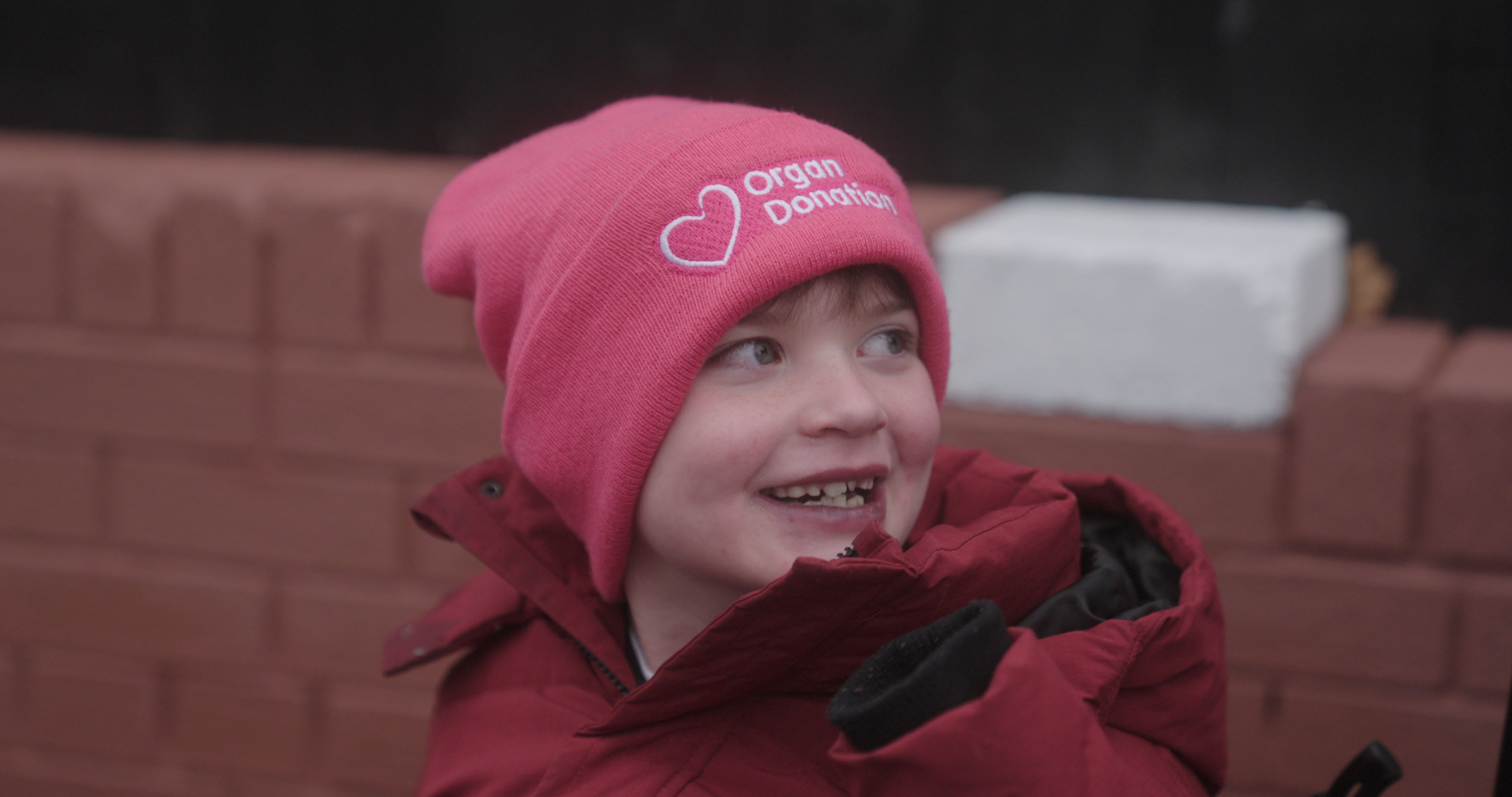 A smiling young boy wearing a pink beanie hat with a 'Organ Donation' message and heart symbol, dressed in a red winter jacket, outdoors near a brick wall.