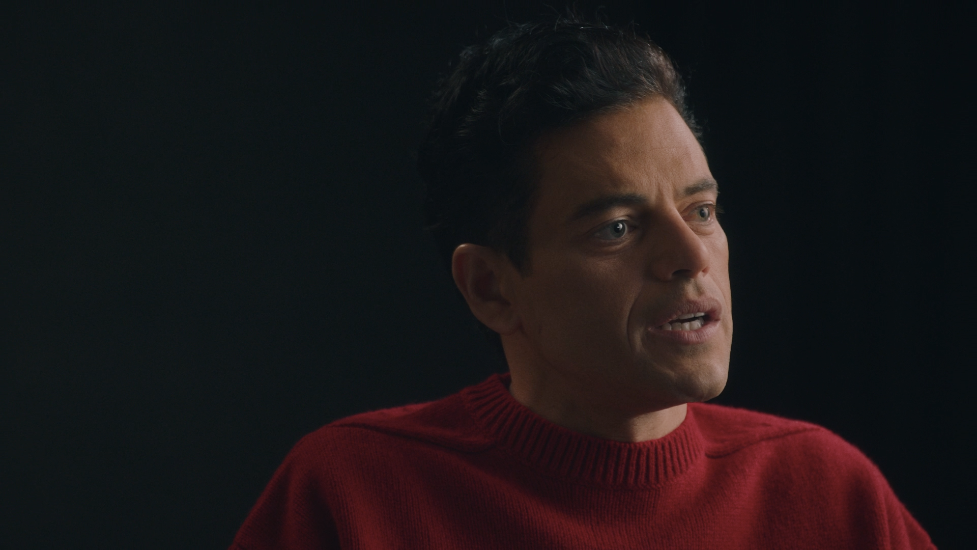 A man with dark hair and blue eyes is wearing a red sweater and looks to the side against a dark background.