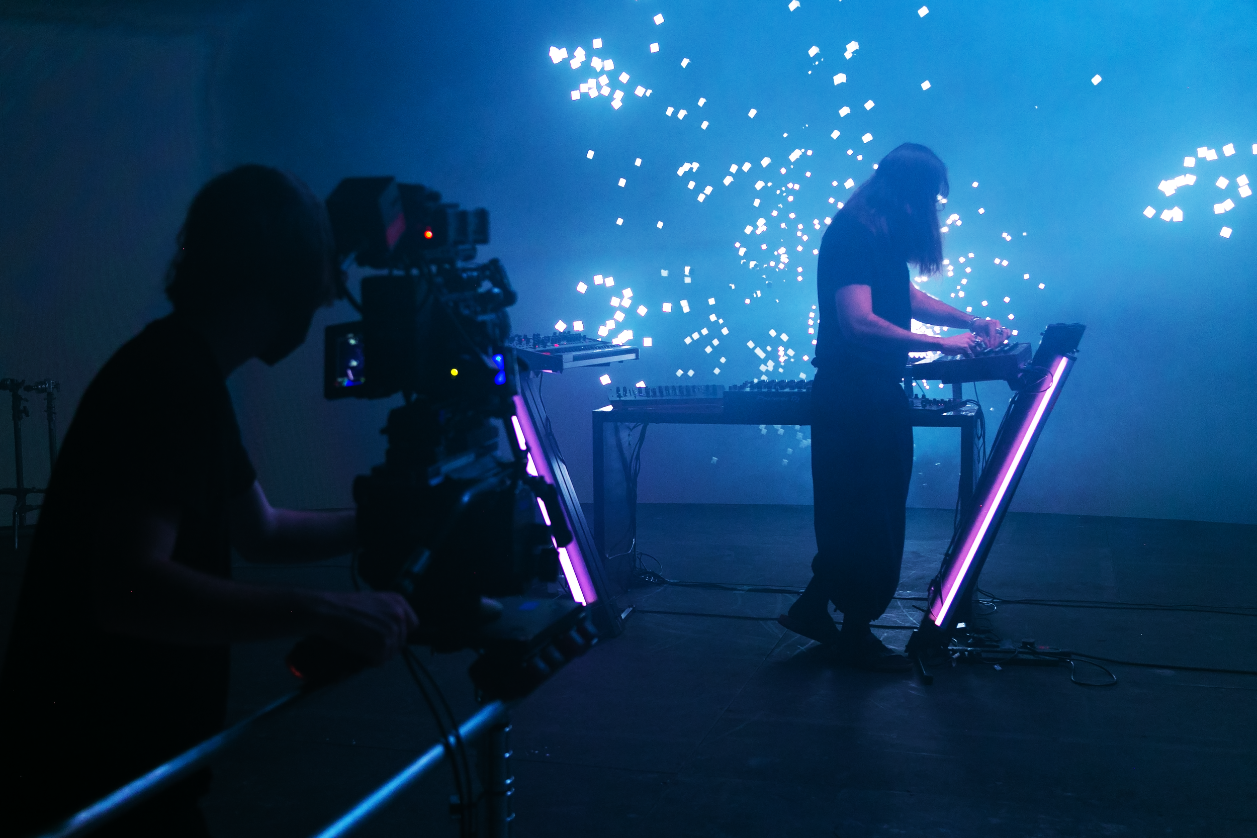 A DJ performs on stage with digital visuals in the background, while a cameraman films the performance in a dark, atmospheric setting.