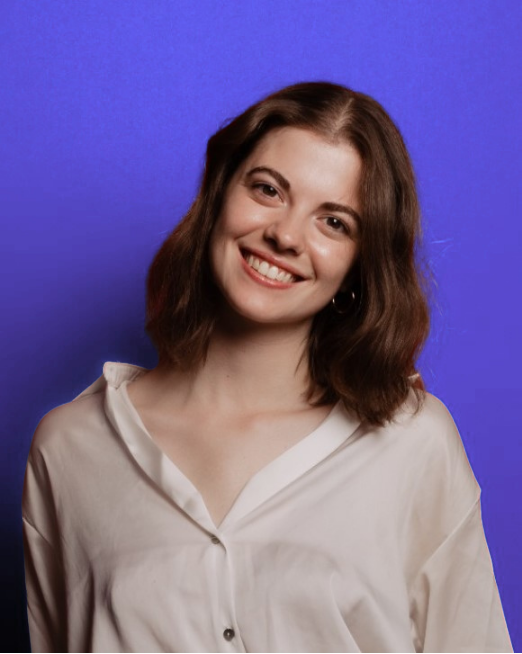 Smiling young woman with brown wavy hair wearing a white button-up shirt against a purple background.