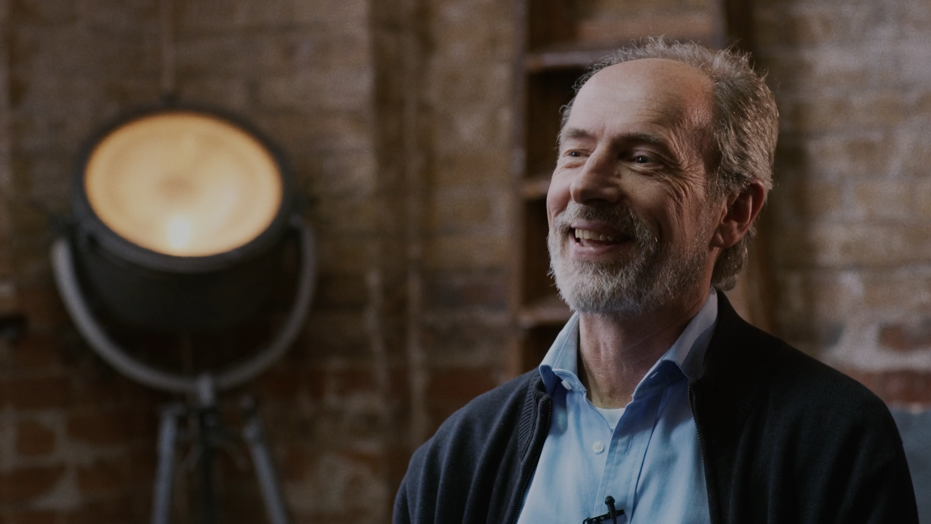 An older man with gray hair and a beard smiling, wearing a light blue shirt and a dark jacket, in a room with brick walls and a large studio light in the background.