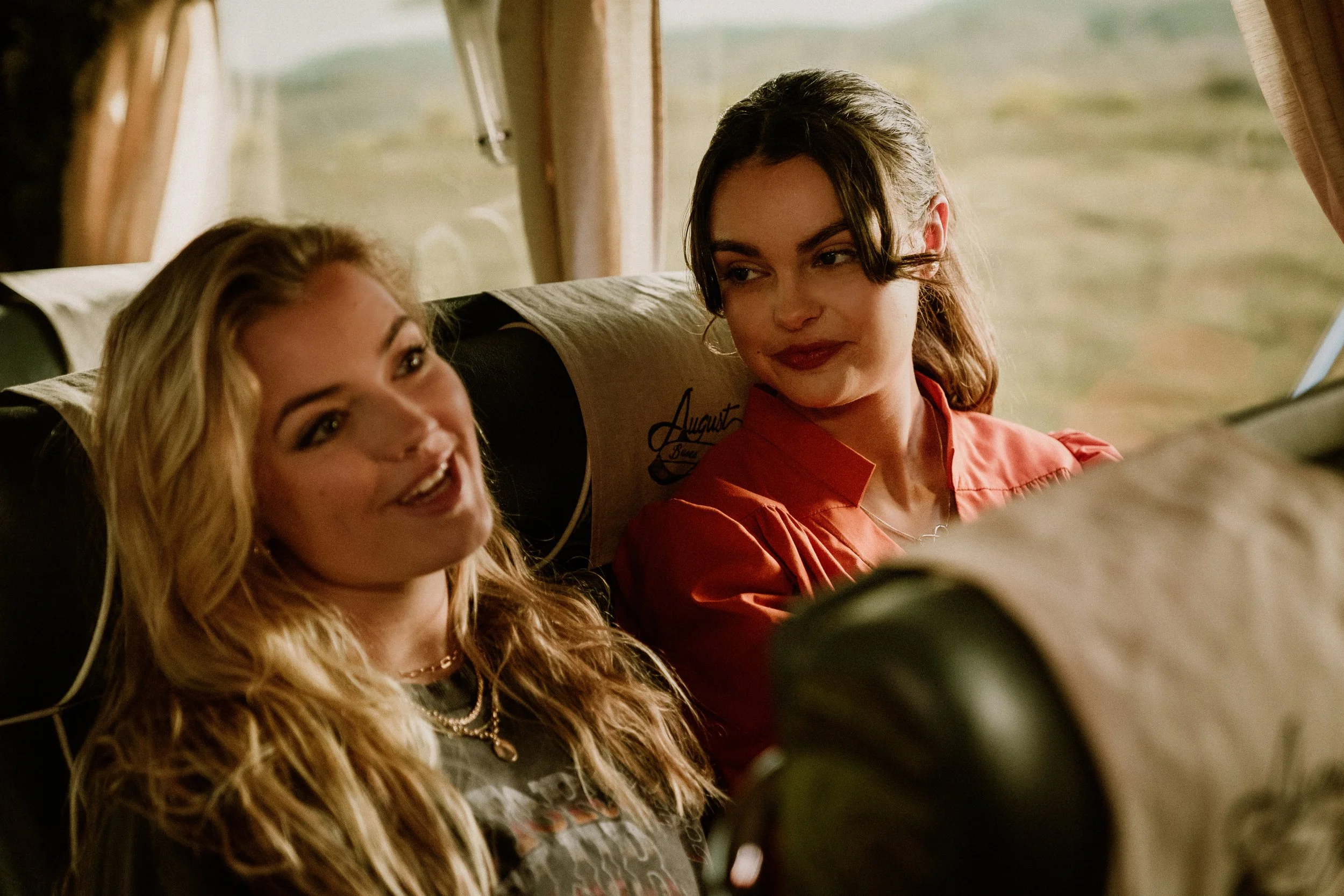 Two women sitting on a bus, one smiling and talking, the other looking at her with a slight smile.