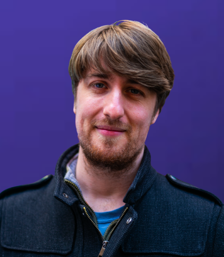 A young man with brown hair and a beard smiling, wearing a dark jacket, against a purple background.