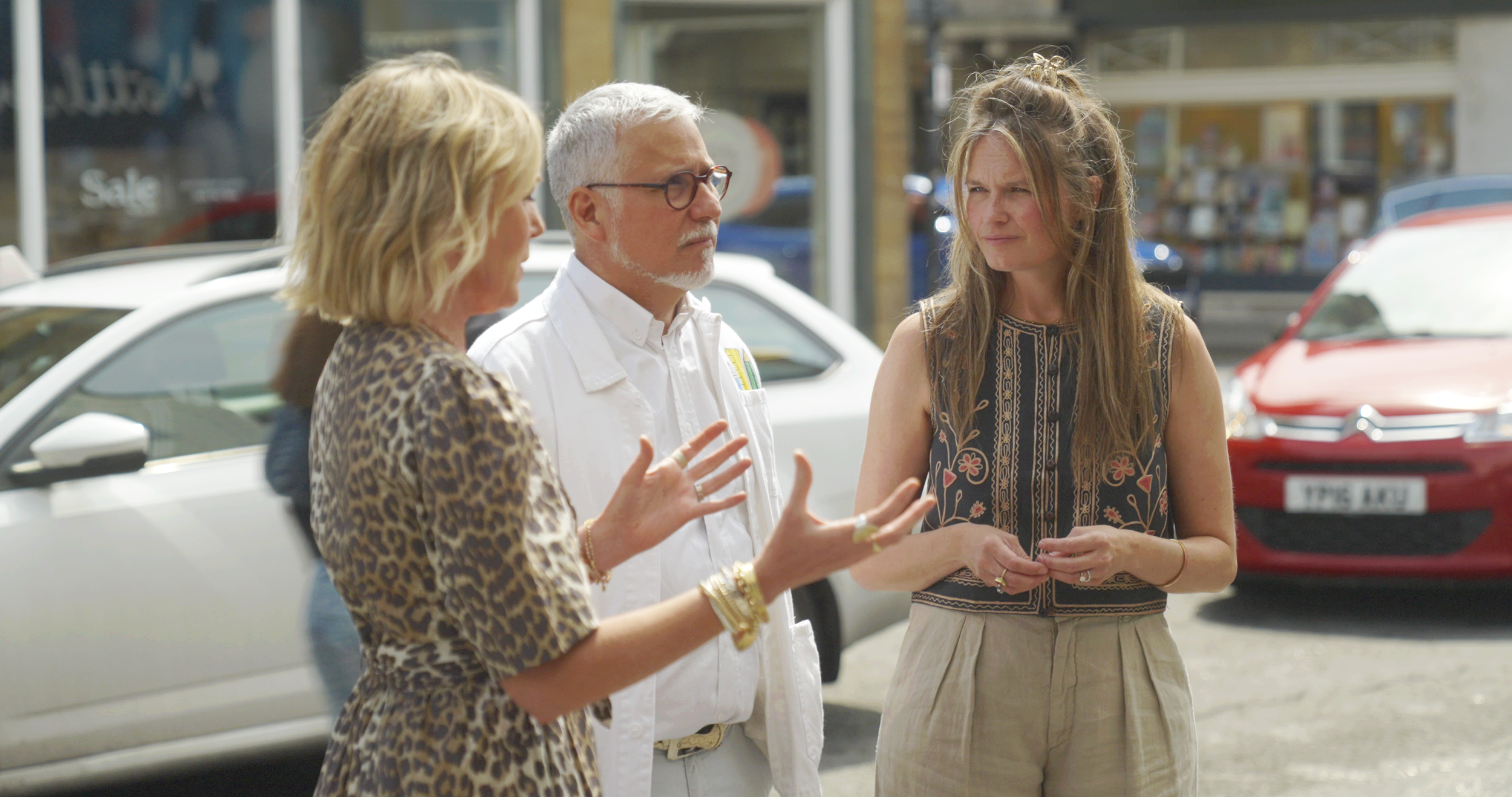 Three people having a conversation outside near parked cars, with a storefront in the background.