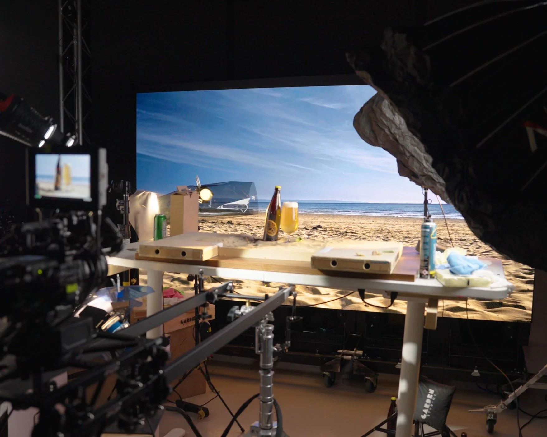 Behind the scenes view of a film or photo shoot set, showcasing a beach scene backdrop, a table with drinks and snacks, and camera equipment.