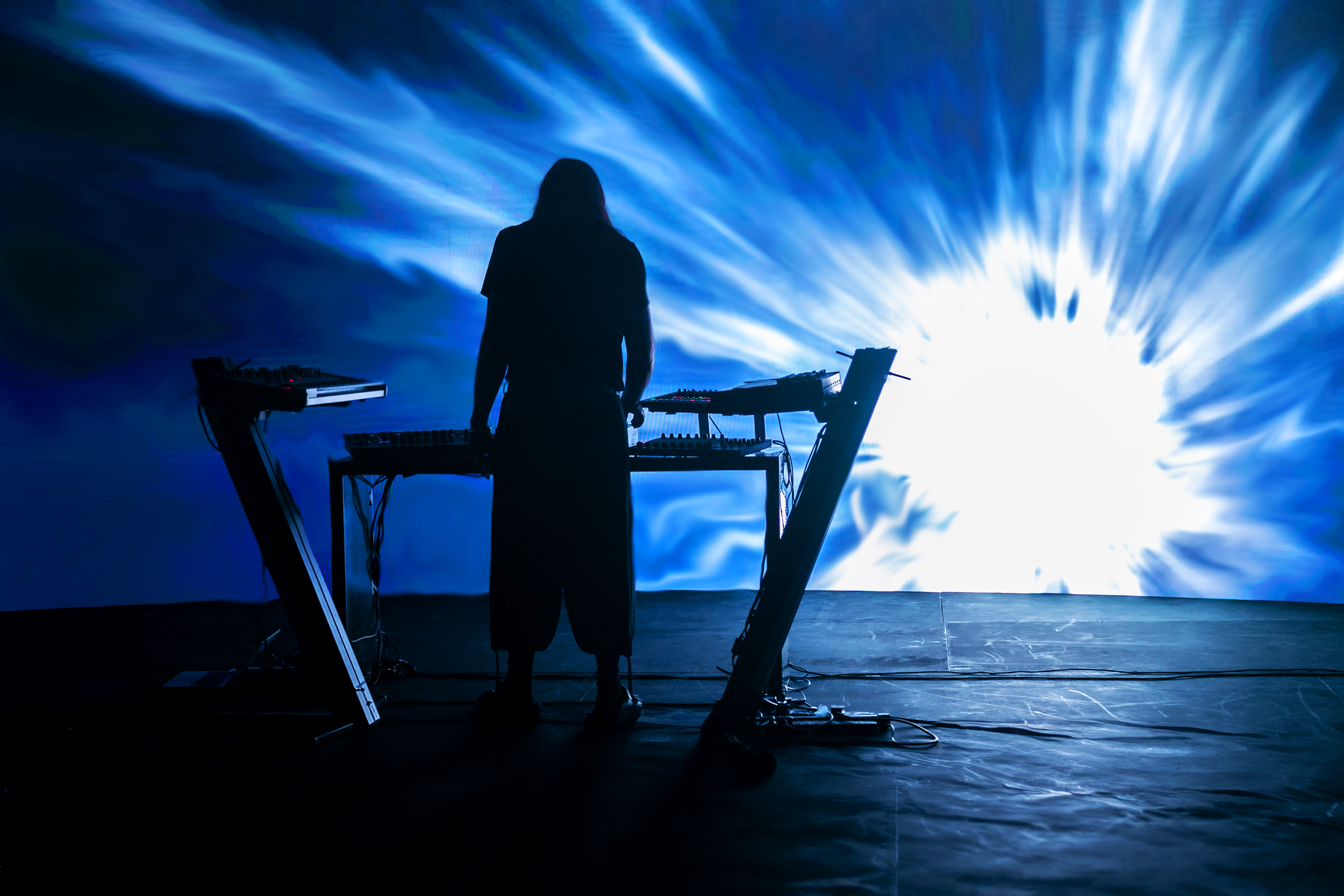 A person silhouetted against a bright, abstract blue and white background, standing behind electronic music equipment such as keyboards and mixers.