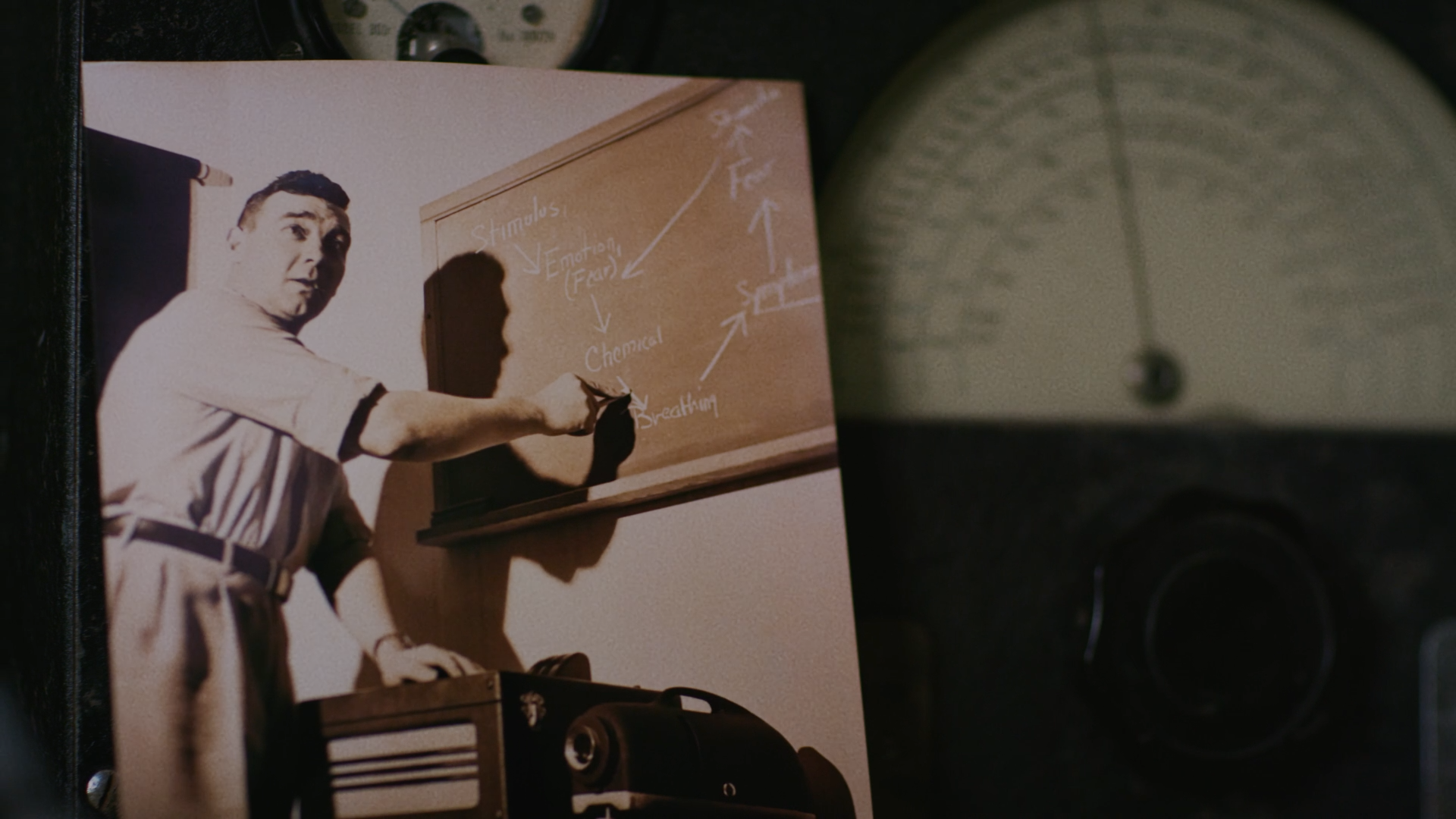 A black-and-white photo of a man pointing at a chalkboard with scientific notes, with a manual typewriter and a desk in the foreground.