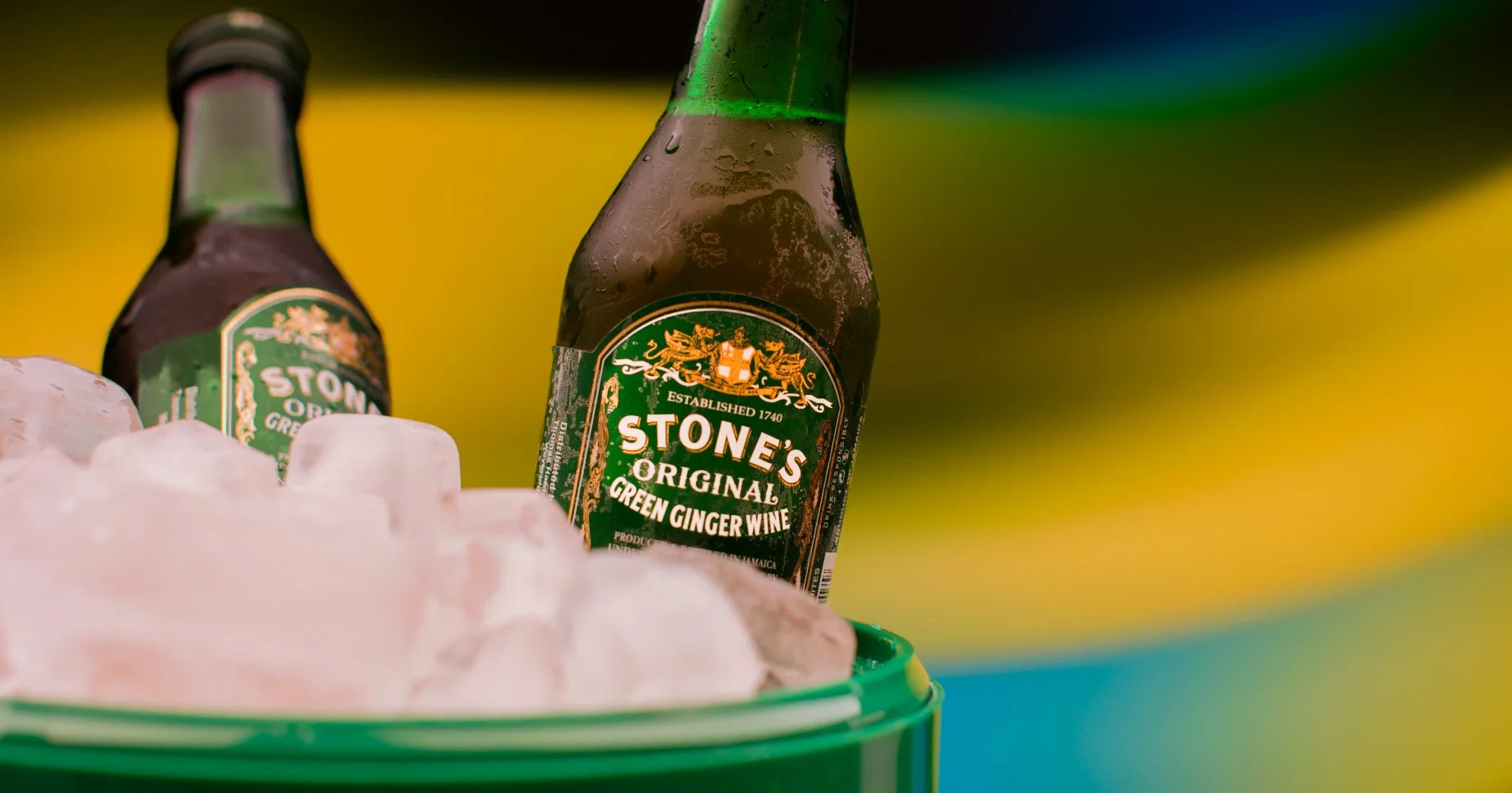 A bottle of Stones Original Green Ginger Wine partially submerged in ice in a green container, with a blurred yellow and dark background.