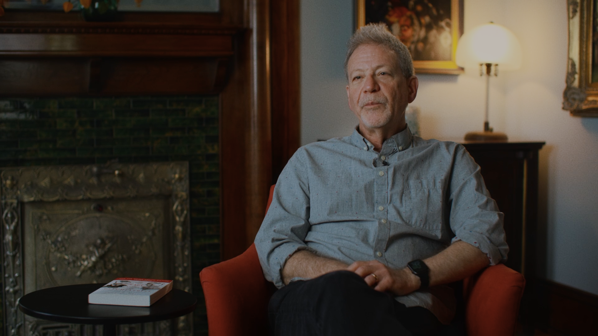An older man with gray hair and a beard sitting in a red armchair in a cozy room with paintings, a lamp, and a fireplace. There is a book on a small black table beside him.