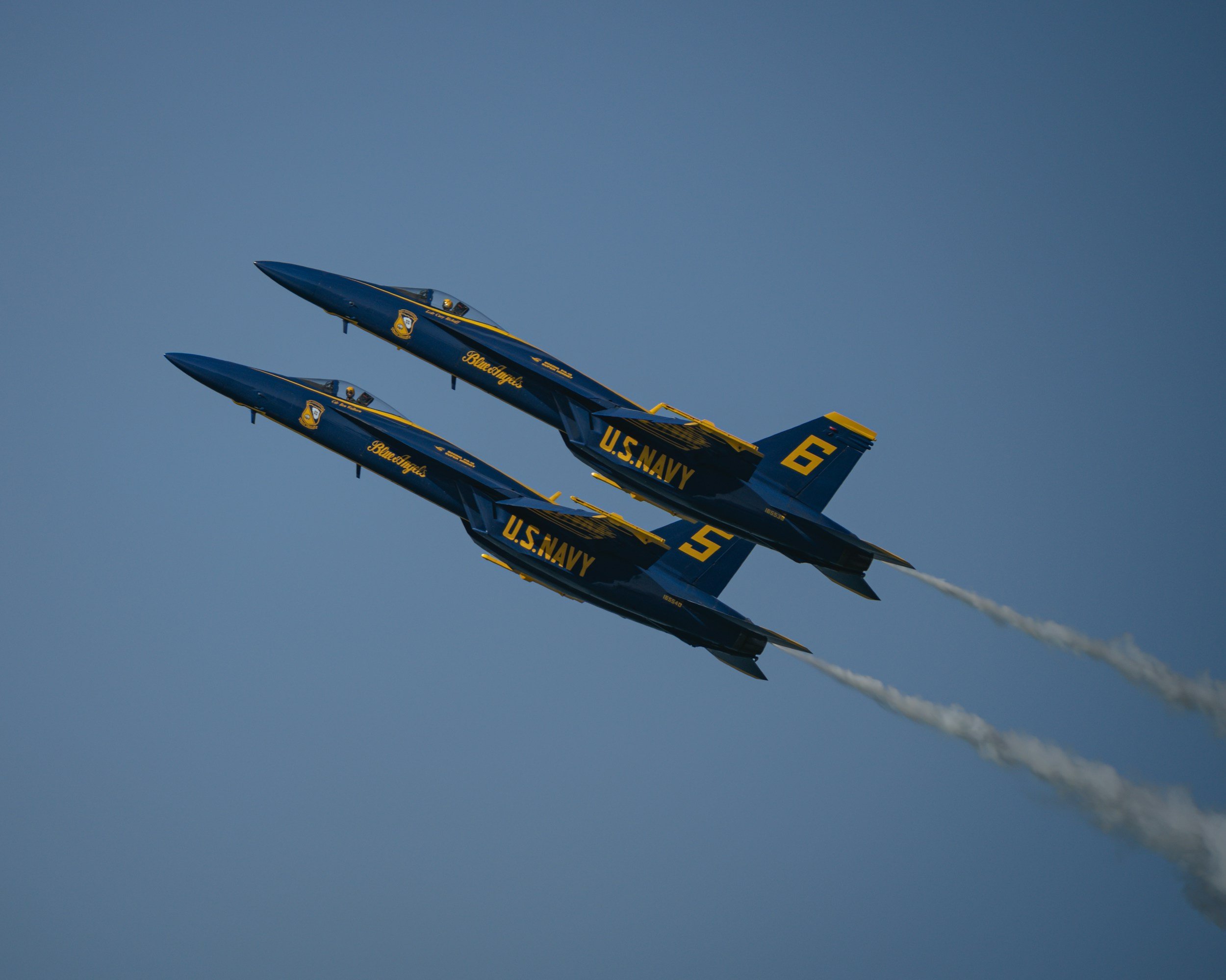 Two U.S. Navy Blue Angels fighter jets flying in tight formation, emitting white smoke trails against a cloudy sky.