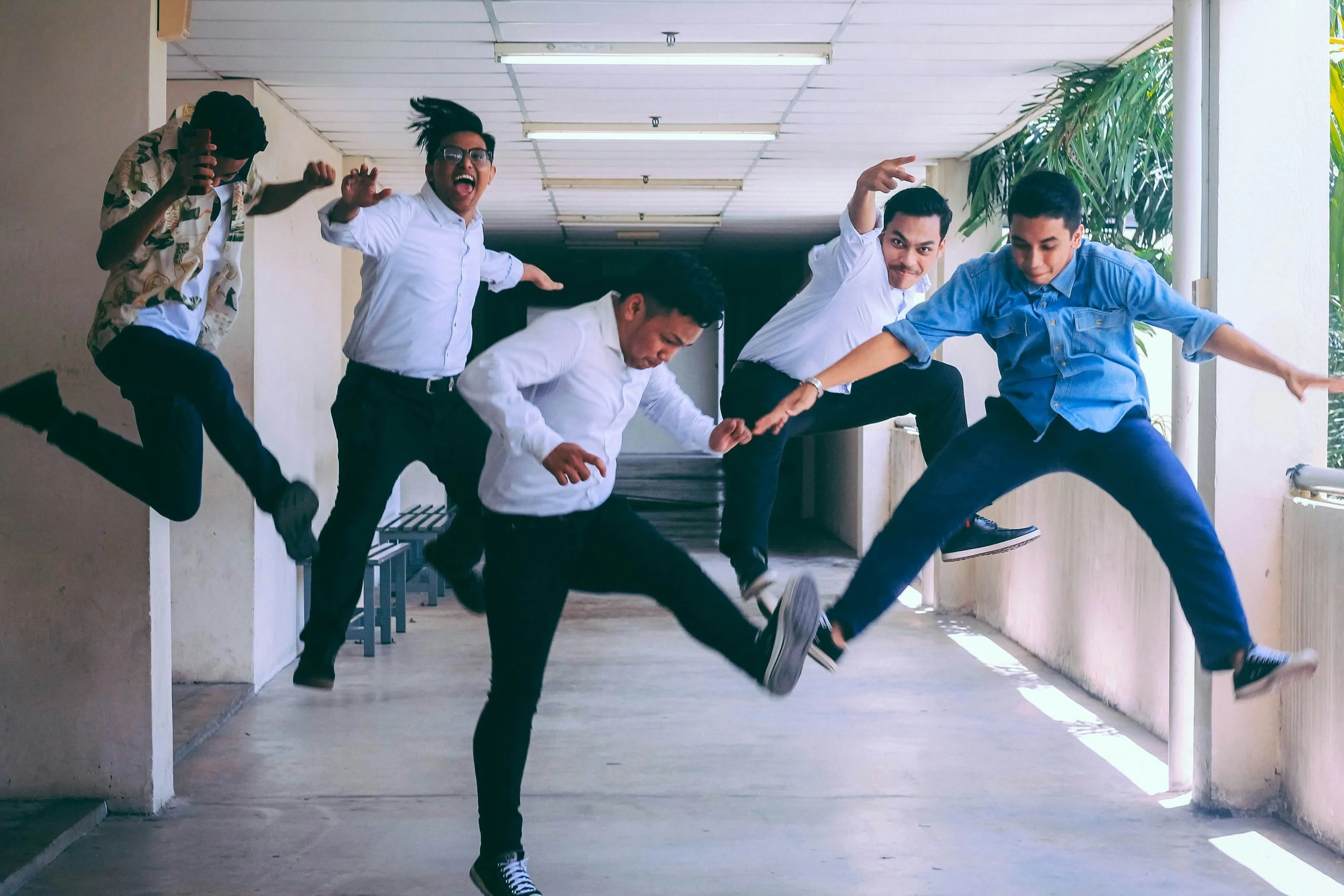Five young people in casual clothing jumping and posing in a corridor with sunlight and greenery outside.