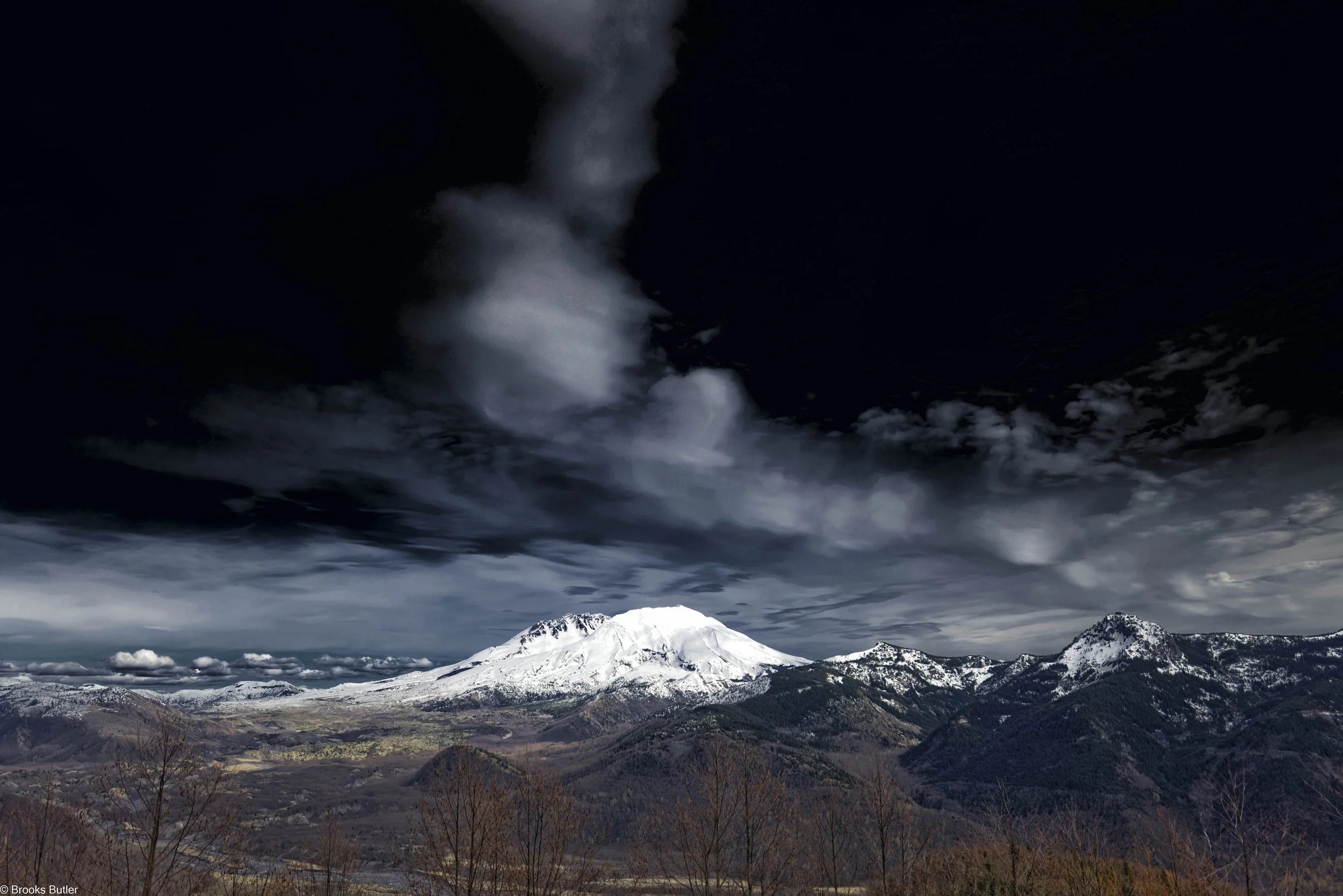 Snow-capped mountains with dark, cloudy skies and mountains in the foreground.