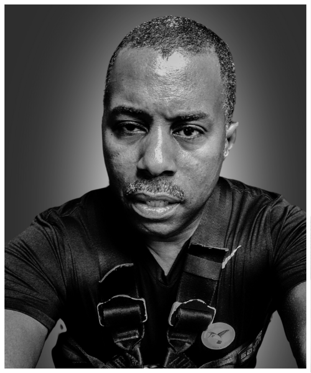 A black and white portrait of a man with short hair, a mustache, and a serious expression, wearing a dark shirt with a badge or button and a strap across his chest.
