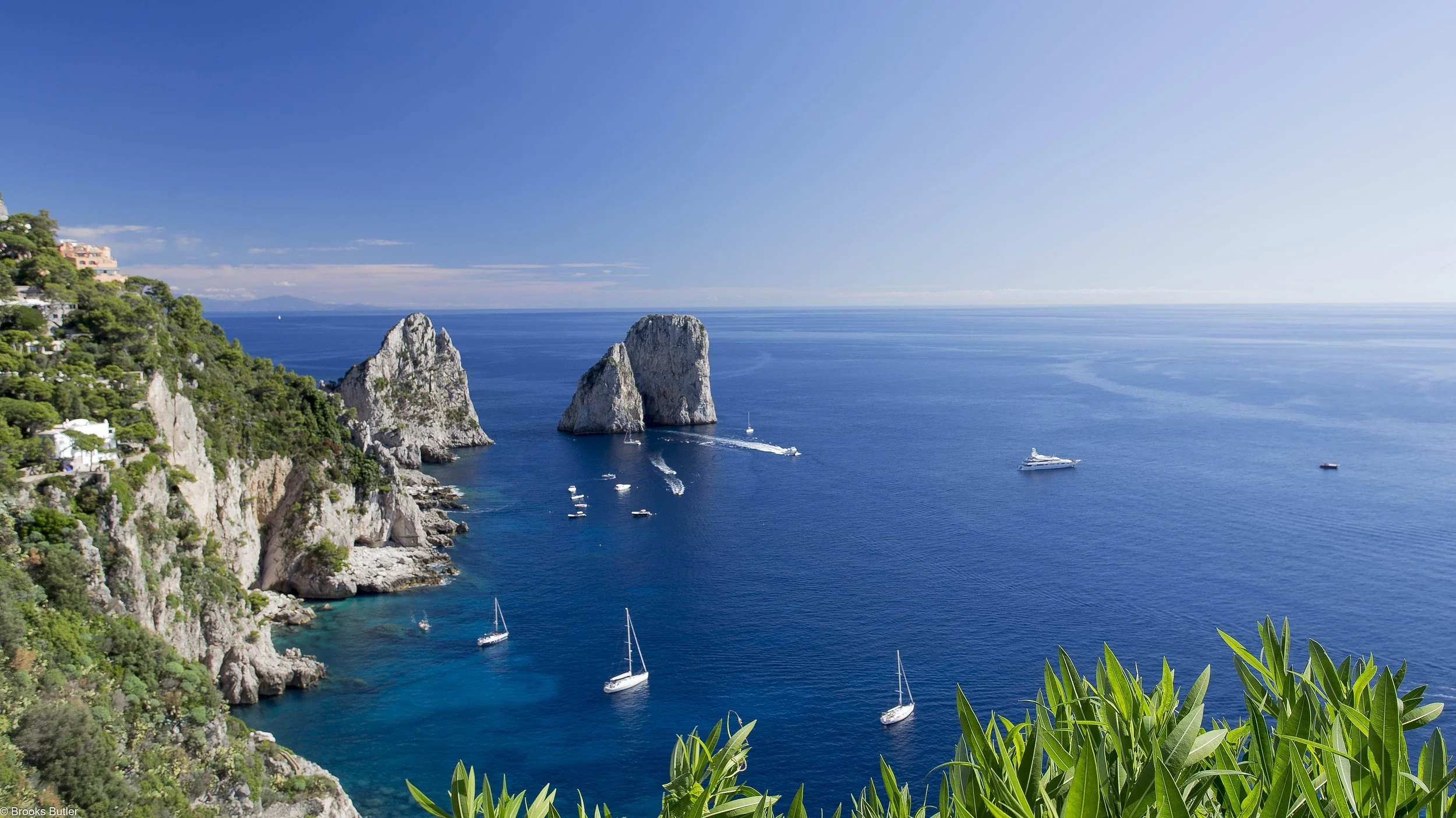 Cliffs with greenery on the left and a view of the ocean with several boats and large rock formations in the water, under a clear blue sky.