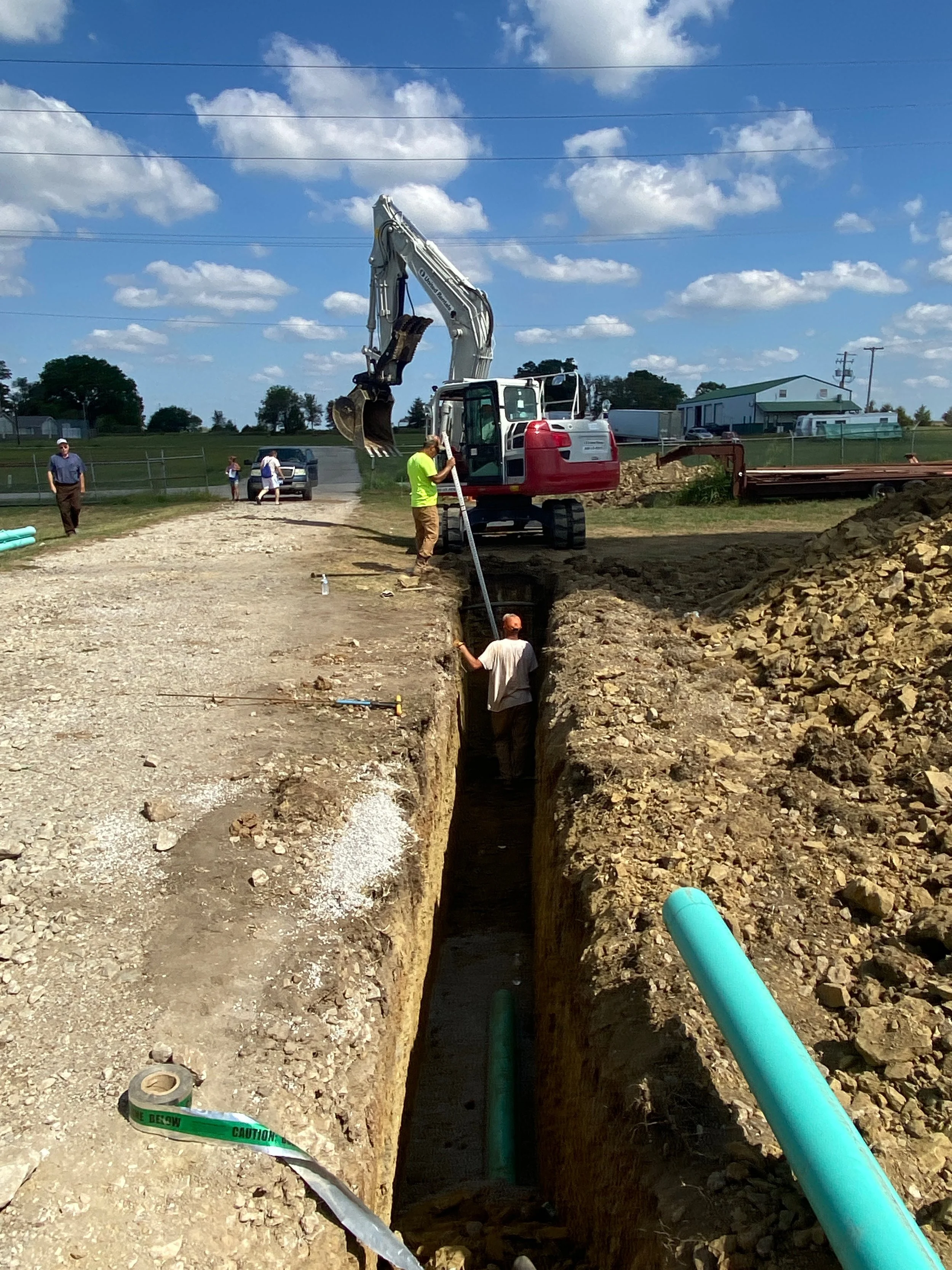 Excavator near trench with workers; construction site with blue sky and clouds.