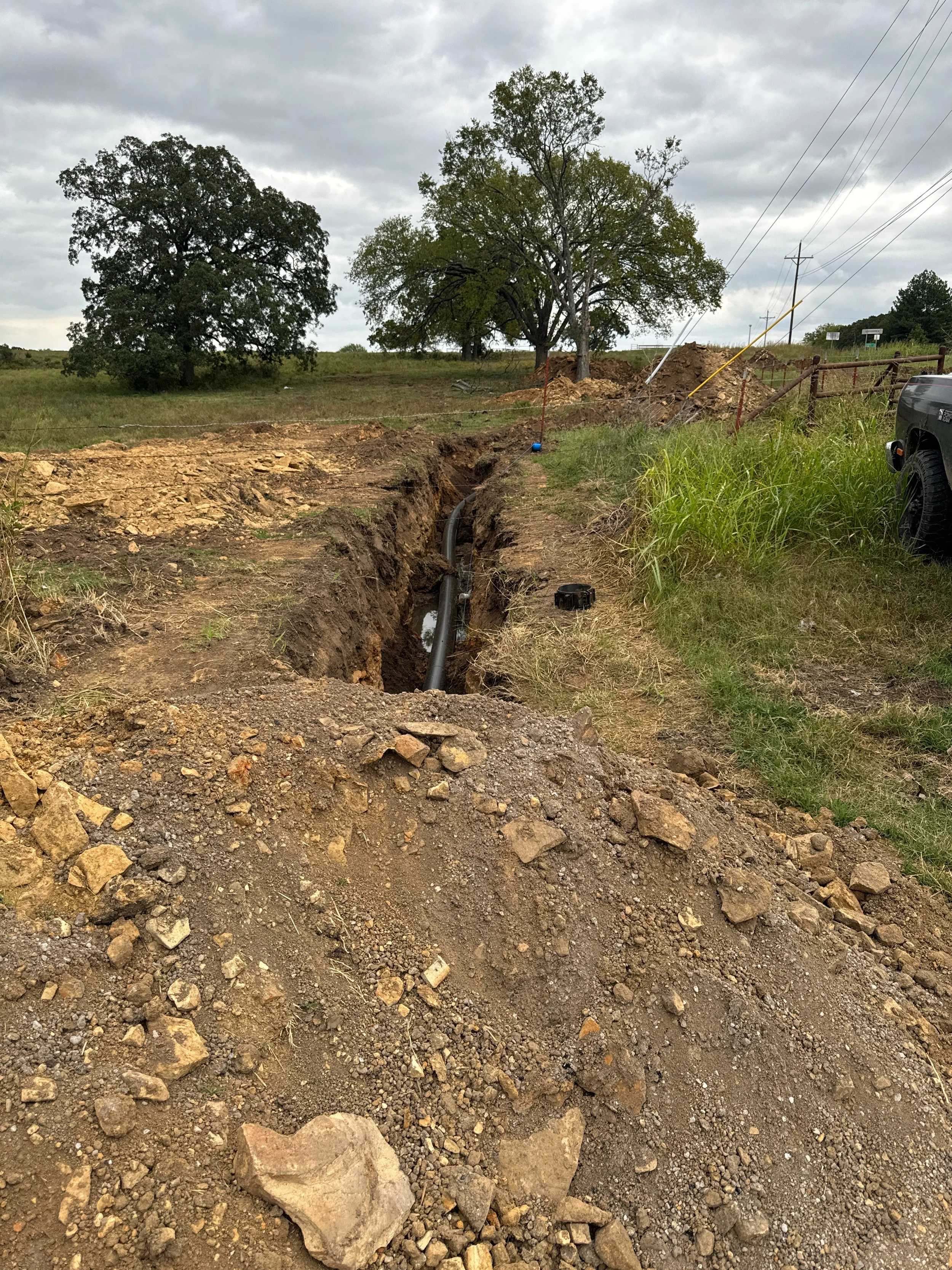 Trench with utility pipe in rural landscape, surrounded by trees and dirt mounds, utility poles visible in background.