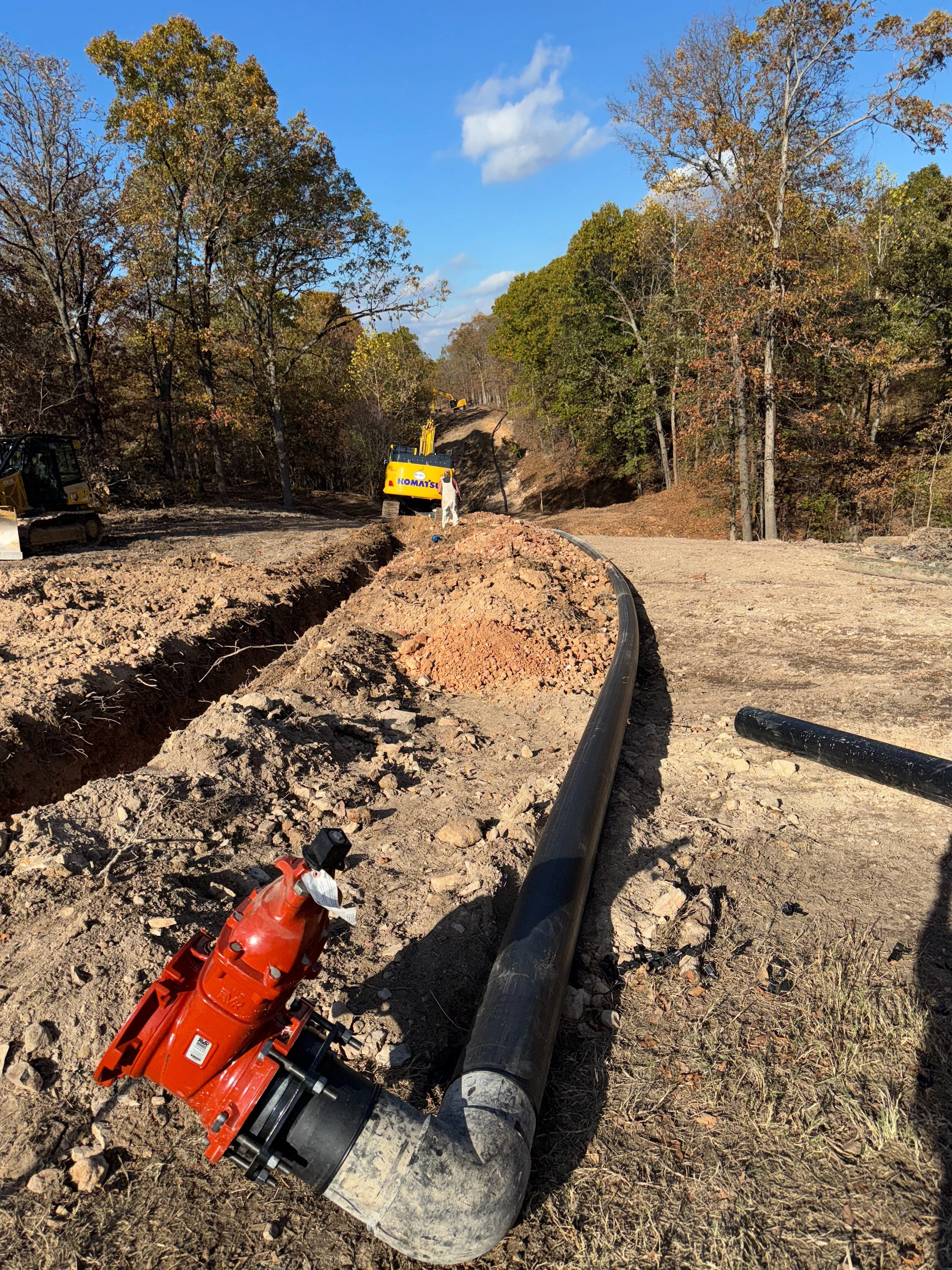 Construction site with a long black pipe alongside a dirt trench, red valve, excavator, and trees in the background.