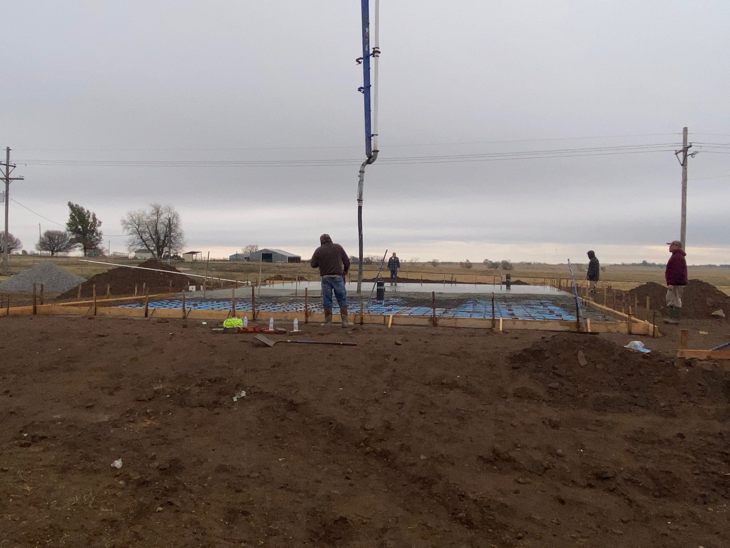 Construction site with workers preparing concrete foundation using a pump truck and wooden formwork, surrounded by dirt and open fields.